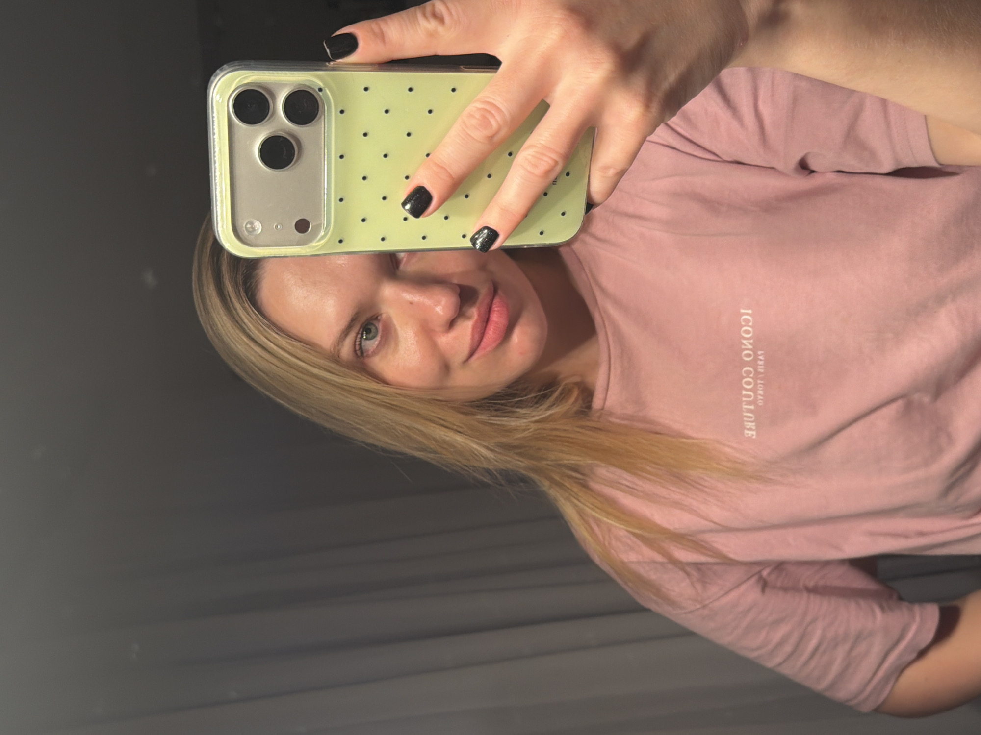 A woman with blonde hair takes a selfie indoors. She is wearing a pink t-shirt with text that appears to say "ICONO COUTURE" printed in reverse. Her nails are painted black. She is holding a light green phone with a black polka dot case. The location is most likely inside a building in Dreiliņi, Latvia.