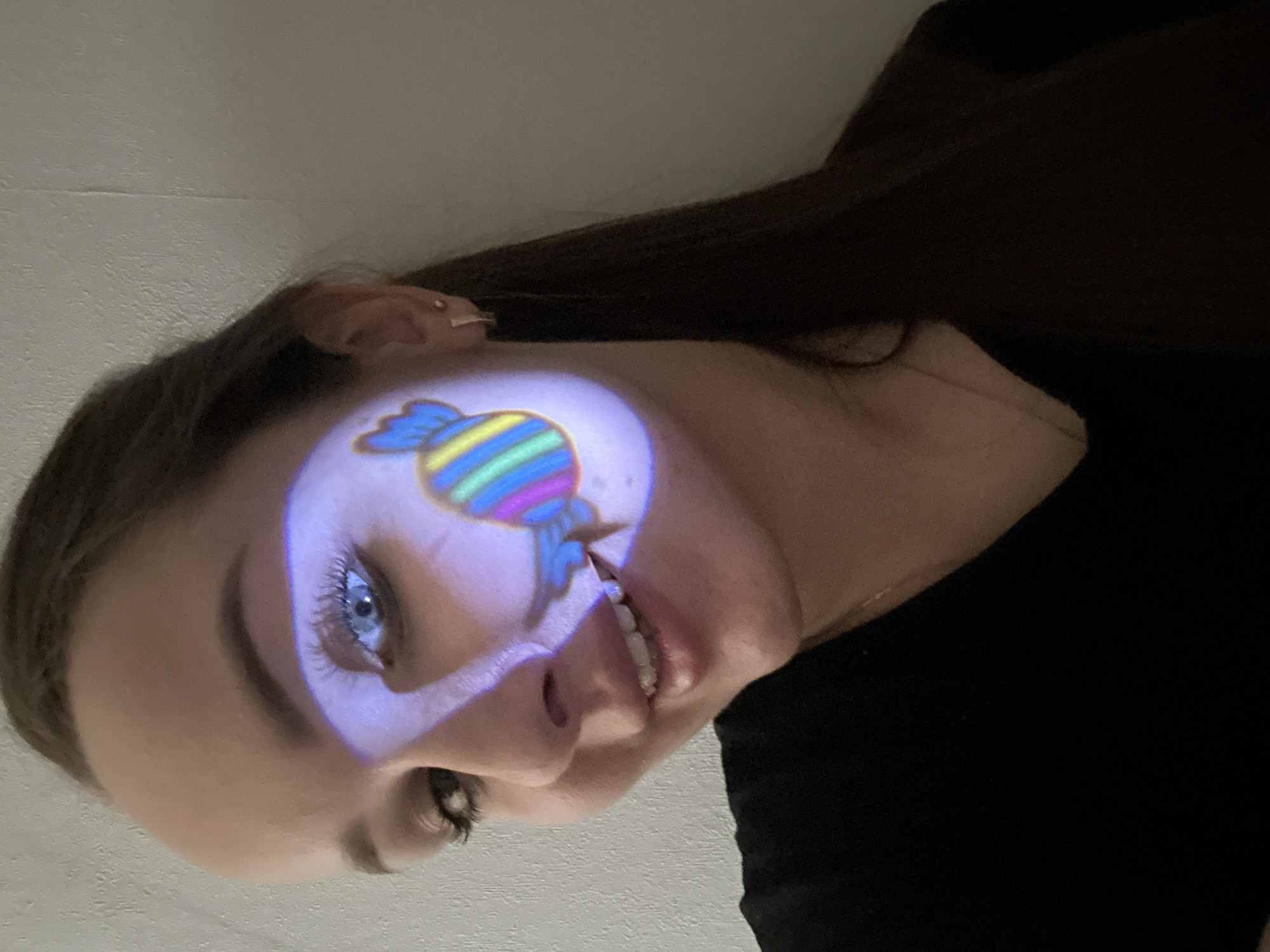 A young woman is prominently featured, likely indoors in Chaikovsky, Russia, with a colorful image projected onto her face. The image is a wrapped candy, possibly a taffy or hard candy, with distinct horizontal stripes in blue, yellow, green, and pink/purple hues. This candy projection creates a soft, luminous purple glow around it on her cheek, extending from below her left eye towards her mouth.
The woman is smiling, showing her teeth, and her bright blue eyes are open and directed slightly upwards. She has long, dark hair that frames her face and flows down, and she is wearing a dark, possibly black, top. A small, light-colored earring is visible in her right ear. The lighting in the scene appears dim, suggesting it might be evening or night, which enhances the visibility of the projected image. The background is a light, neutral color, resembling a wall or ceiling. The overall impression is one of playfulness and cheerfulness. There is no visible text in the image.