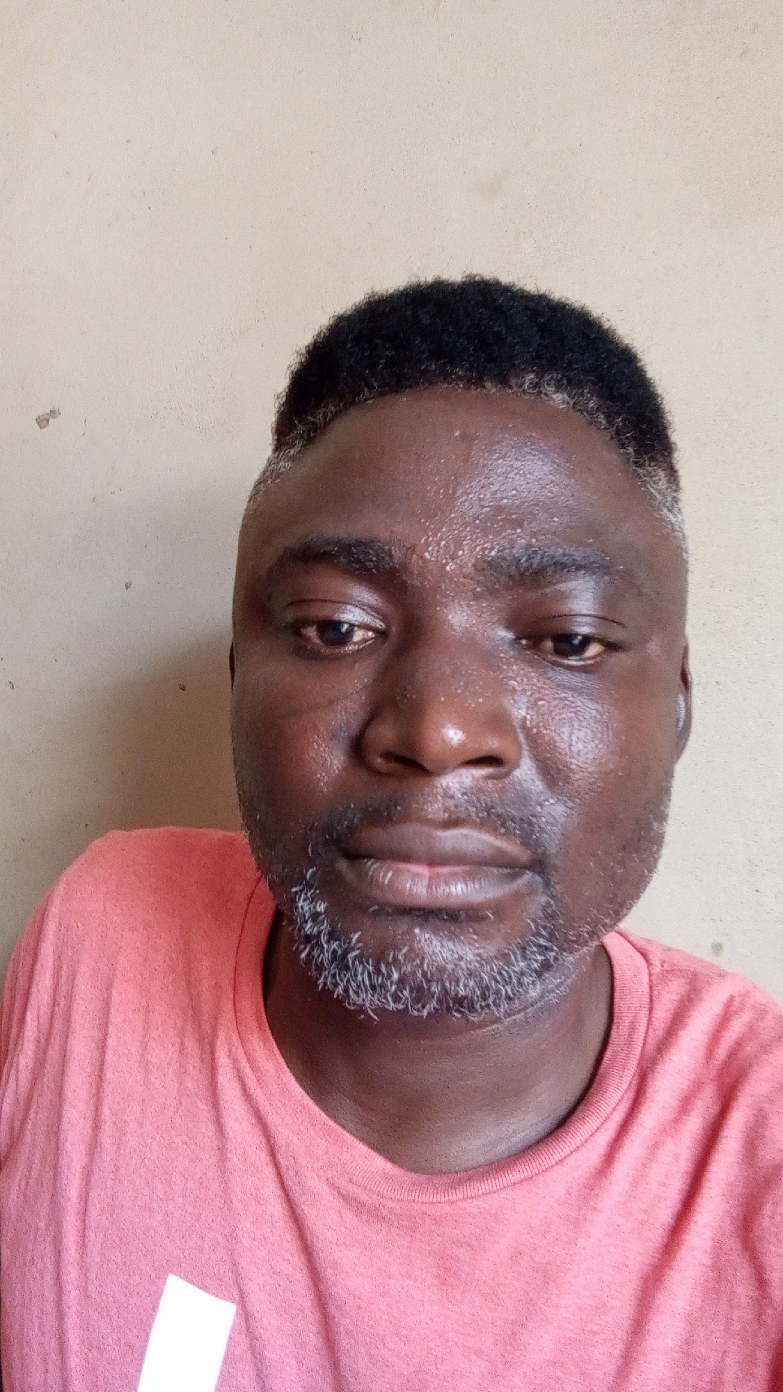 A medium close-up portrait shows an adult Black man looking directly at the camera. He has dark skin with a noticeable sheen on his forehead, nose, and cheeks, suggesting sweat or oiliness. His hair is dark and curly on top, with visibly graying, closely cropped sides and temples. He has a full beard and mustache, which are predominantly gray. A faint scar is visible on his right cheek, below his eye. The man is wearing a short-sleeved, crew-neck t-shirt in a salmon or light coral color. A partial white geometric shape, possibly a logo or design, is visible on the lower left side of his shirt. His expression is neutral. The background is a plain, light beige or off-white wall with a slightly textured surface and a few minor imperfections. The lighting appears even, suggesting indoor conditions or an overcast day.