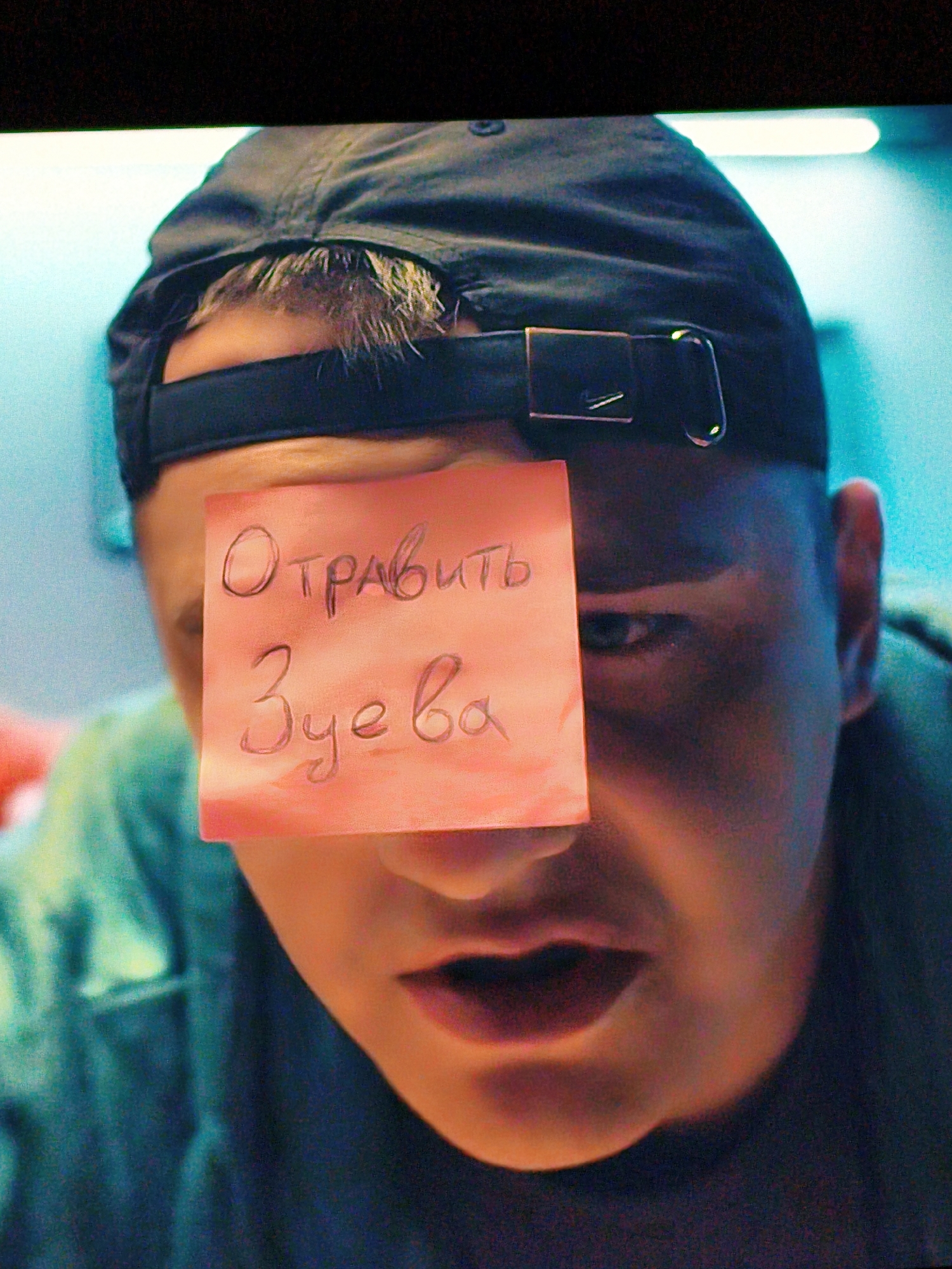 A person wearing a black baseball cap has a pink sticky note covering part of their face, obscuring their right eye and nose. Handwritten text in Cyrillic script is visible on the sticky note. The text appears to read "Отравить Зуева" which translates to "Poison Zuev". The person is looking slightly downwards and to the left. Their mouth is slightly open, and they appear to be wearing a teal garment. The background is a blurred mix of teal and dark tones, suggesting an indoor environment. No specific location in Vladivostok, Russia is identifiable.