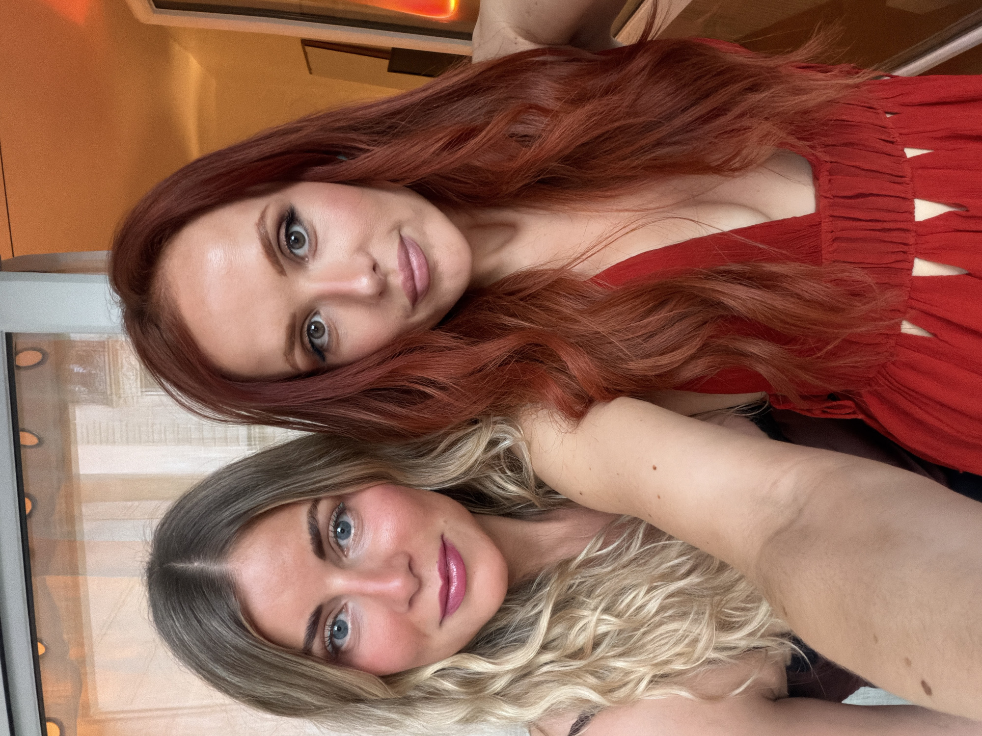 Two women are posing close together for a selfie indoors, likely at a high vantage point in Paris, France. The woman at the top of the frame has red, wavy hair and is wearing a red dress. She has light skin, and dark eye makeup. The woman at the bottom of the frame has blonde, wavy hair. Both women have light skin and are wearing makeup, including lipstick. The background includes a window with a view that suggests a high elevation within the city. There is a warm lighting tone in the image.