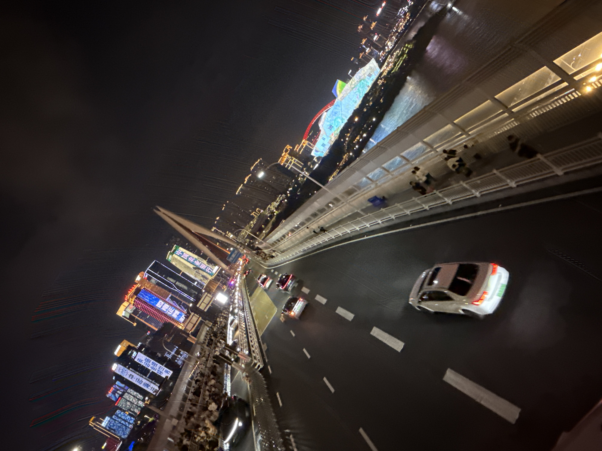 This image captures a nighttime view of the Yuzhong District in China, likely from a bridge or elevated vantage point. The scene is dominated by the illuminated cityscape, with numerous tall buildings adorned with vibrant lights and large digital displays. A bridge, likely a cable-stayed bridge, is prominently featured, with its suspension cables visible against the dark sky. A road runs across the bridge, with several cars visible. One white sedan is in the foreground, its taillights illuminated. On the walkway of the bridge, several pedestrians are visible, some appearing to be standing and observing the scenery, while others are walking. The river below the bridge reflects the city lights, adding to the dynamic and colorful atmosphere of the scene. The overall impression is of a bustling urban environment at night, characterized by striking architectural lighting and a significant amount of human activity.