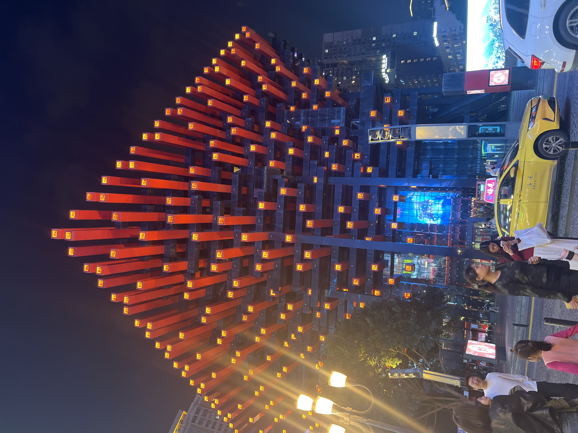 The image shows the Raffles City Chongqing complex located in Yuzhong District, China, at night. The most prominent feature is a large architectural structure composed of red and dark gray cantilevered elements with illuminated square insets. The structure is diagonally oriented in the frame, appearing as a multifaceted cube. Below the structure are street lamps, and several people are standing on the sidewalk looking at the camera. Beside the structure are buildings with digital screens displaying blue light, as well as storefronts. A yellow taxi cab is parked along the curb. In the upper-right corner, part of a white vehicle is visible. The sky is dark.