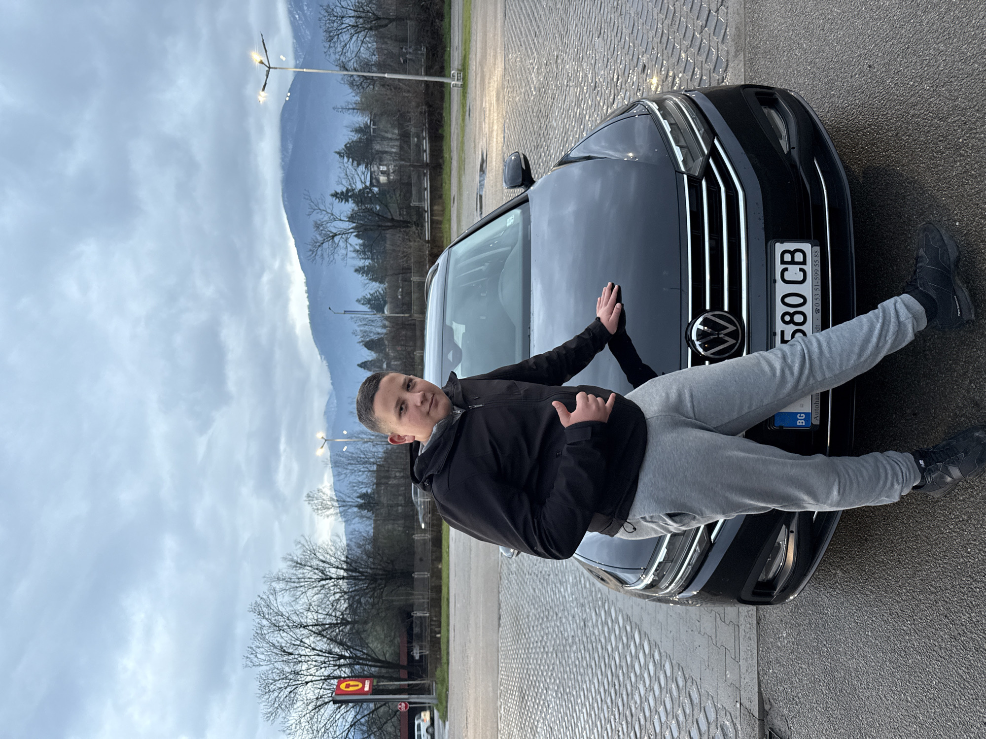 A young man is reclining on the hood of a black Volkswagen car with license plate number "580 CB," likely in the city of Berkovitsa, Bulgaria, given the Bulgarian license plate. He is wearing a black jacket and grey sweatpants and black sneakers. He is making a hand gesture. Behind him is a mountainous landscape under a cloudy sky. A streetlamp and bare trees are also visible. The car is parked on a paved surface. A red and white sign is partially visible at the bottom left.