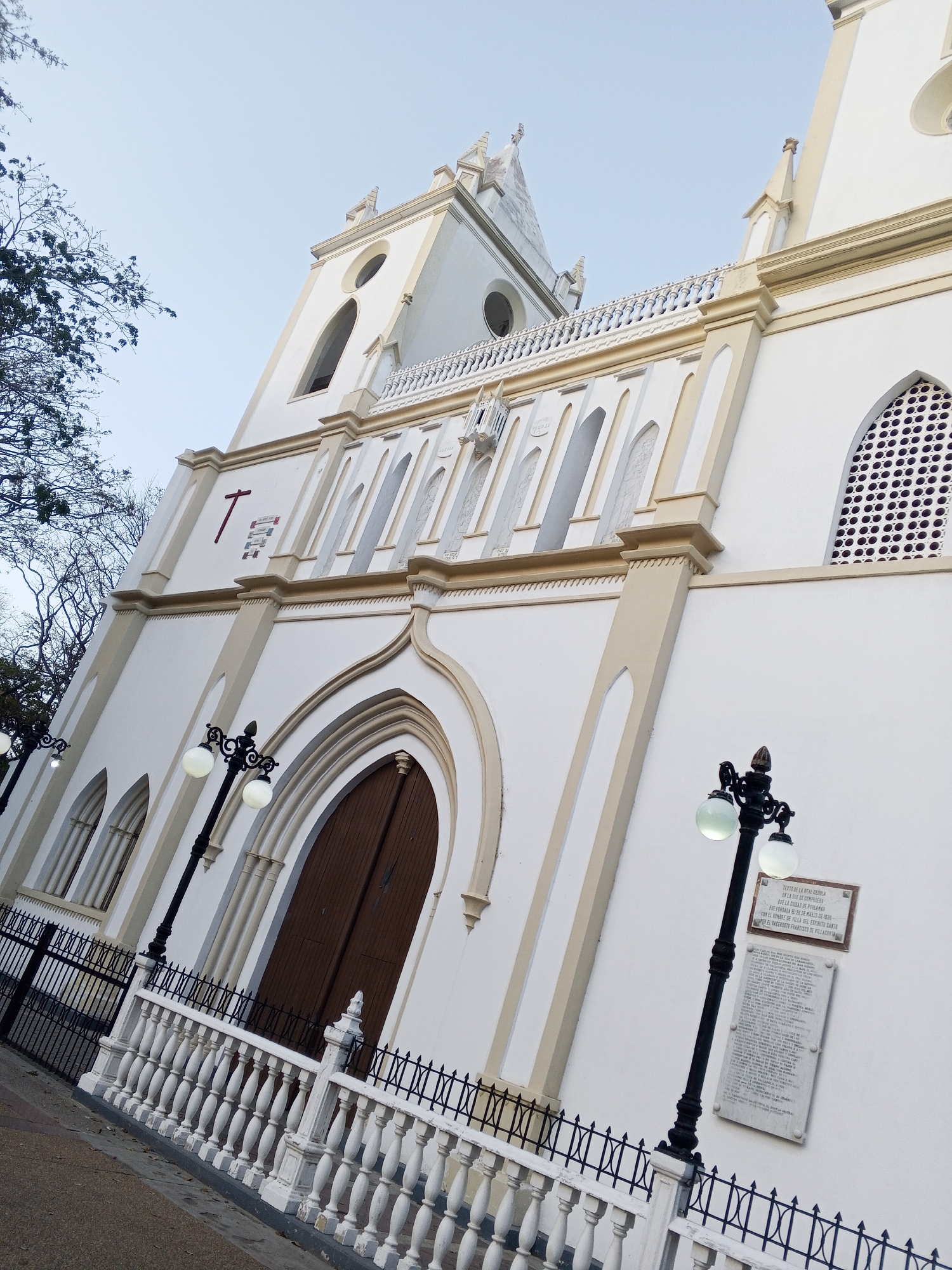 The image shows a low-angle shot of a white and beige building, possibly a church or religious structure, under a bright sky. The building has a tower, arched windows, and a large wooden door. A white balustrade fence with a black wrought-iron fence behind it runs along the base of the building. Two ornate black lampposts with spherical white lamps are positioned in front of the building, and a plaque with text in Spanish is visible on the wall next to one of the lampposts. Bare tree branches are visible in the top left corner of the image.