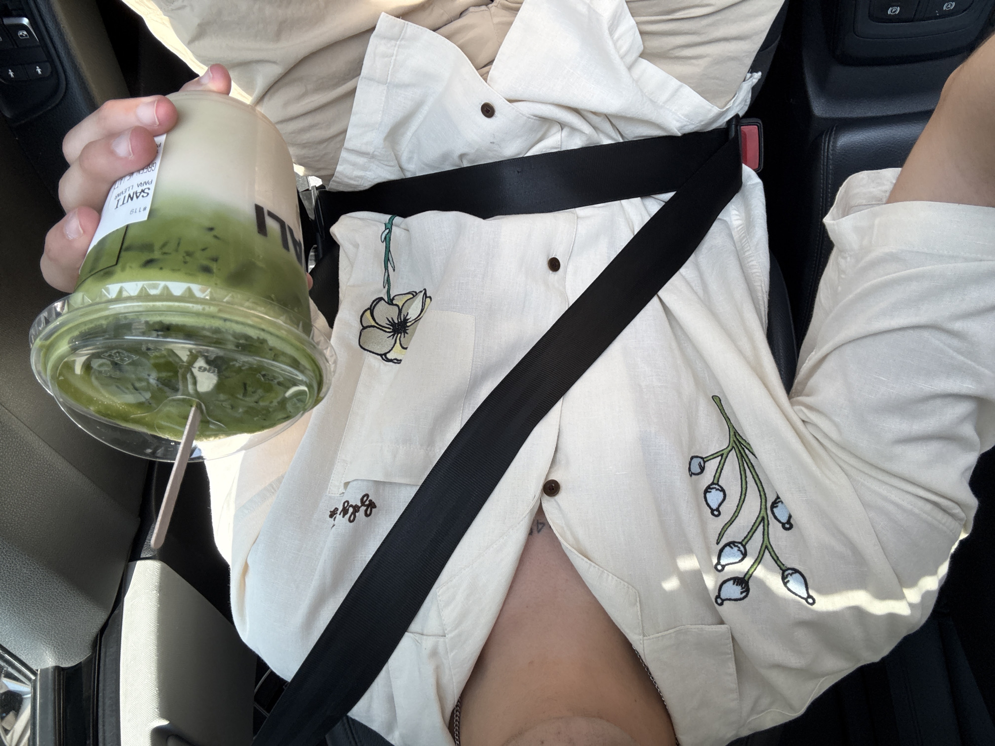 The image shows a person in the passenger seat of a car wearing a light-colored button-down shirt with floral embroidery and a seatbelt fastened across their body. The person is holding a plastic cup filled with a green drink, possibly matcha latte, with a straw. Part of the car's interior is visible in the background, with the console and part of the driver's side. Text on the cup reads "SANTI" and "#119 GREEN VE LATT PARA LLEVA" and "Bologa". Based on the text "SANTI" and the phrase "PARA LLEVA" which is often associated with "para llevar" or "to go", and Bolgá, it is suggested that this image was likely taken in Puebla, Mexico, where this establishment is located.