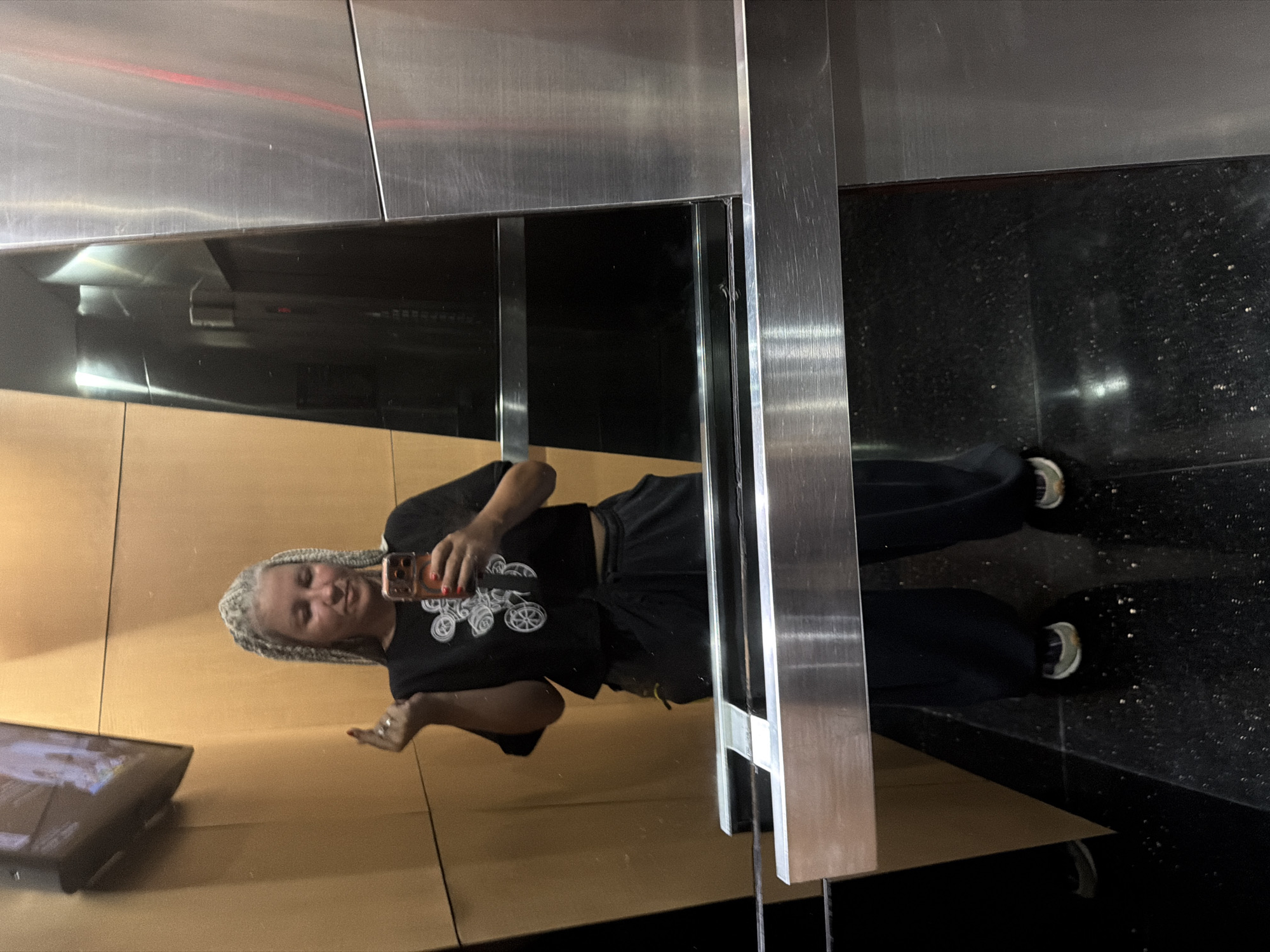 The image depicts a reflection in a mirrored surface, possibly in South Jakarta, Indonesia, although this cannot be definitively confirmed from the image alone. A person with light-colored braided hair is holding a mobile phone and taking a selfie. The person is wearing a dark t-shirt with a graphic design and dark pants. The reflection shows part of a digital display, likely a screen, and a metallic structure in the foreground. The floor near the mirrored surface is dark and appears to have a speckled pattern. The overall impression is that the person is in a commercial or public space with modern architectural elements.