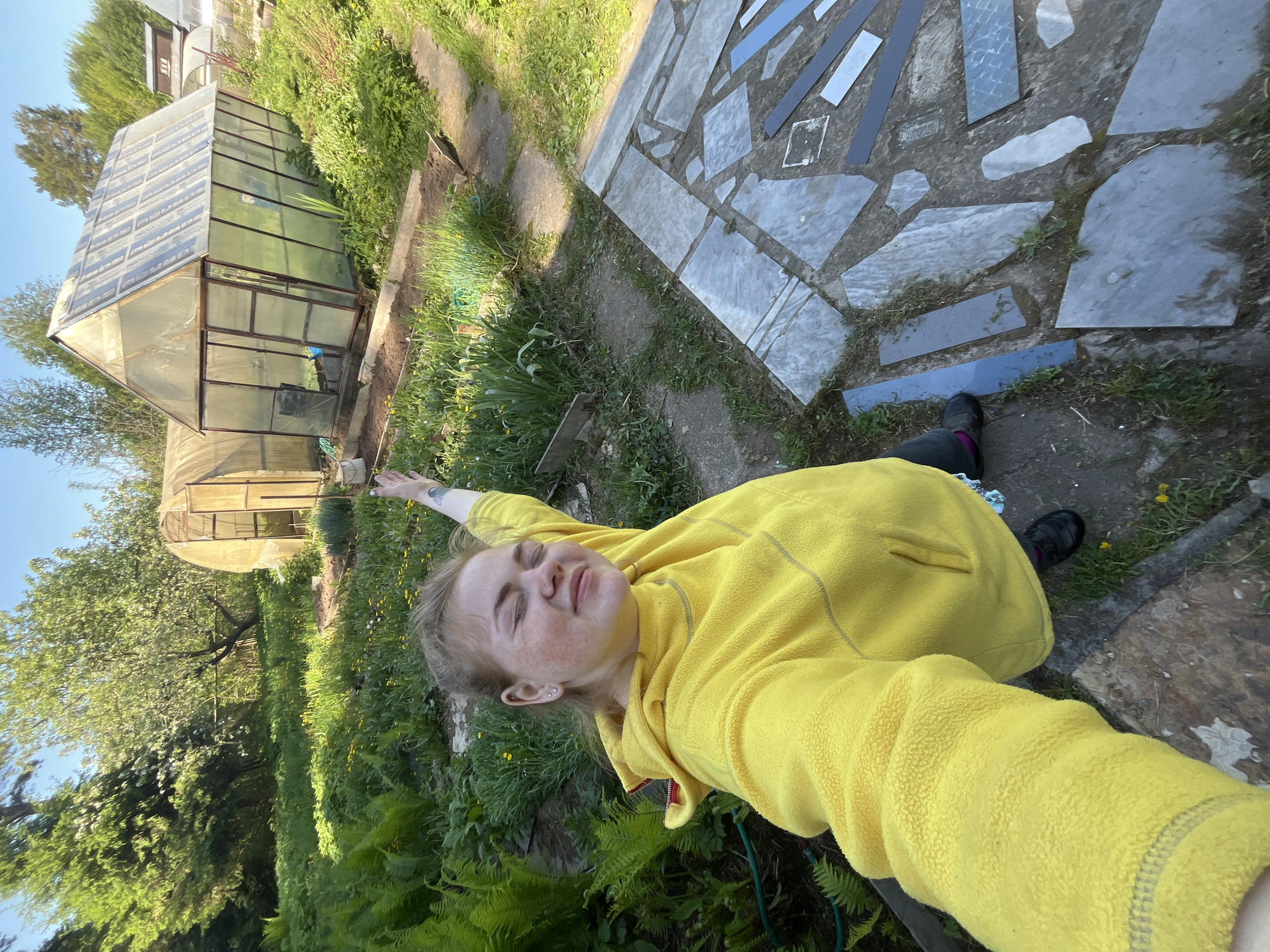 A woman in a yellow fleece jacket lies on a concrete walkway. The walkway is decorated with grey stones and blue glass fragments. To the left of the walkway is a green lawn with ferns. Beyond the lawn is a greenhouse with a metal frame and glass panels. The location is Chaikovsky, Russia.
