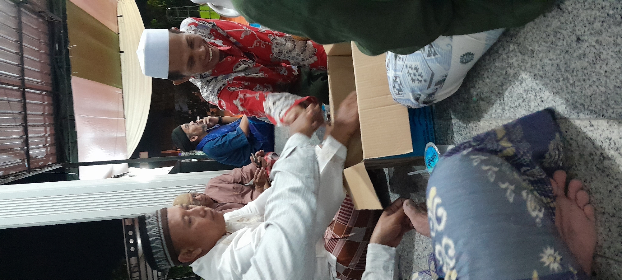 In Bendungan, Indonesia, a group of men are seated on the ground, likely in a communal gathering or religious event. In the foreground, one man in a white shirt and patterned sarong is shaking hands with another man wearing a red shirt with a white pattern and a white cap. To the left, another man in a light brown shirt and glasses is looking down at his phone. Further back, a man in a blue shirt and black cap is seen holding a cigarette and speaking on a phone. A cardboard box is placed on the ground in front of the men, with a small blue cup nearby. The setting appears to be outdoors, under a covered structure with striped fabric hanging above, suggesting it might be an event space or a shaded outdoor area. The ground is a rough, grey textured surface.