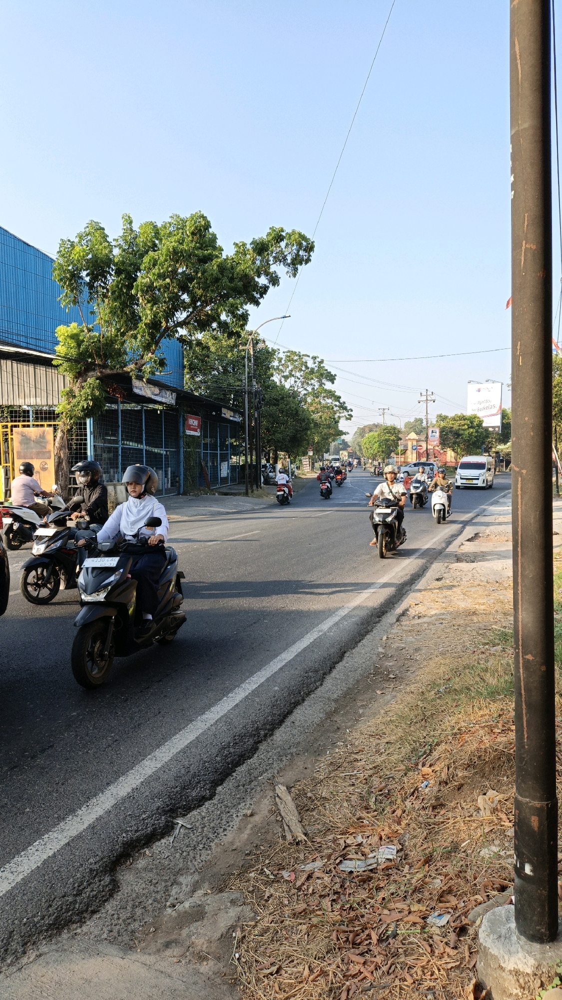The image depicts a street scene in Kuwasen, Indonesia, under a clear, bright sky. A busy road is filled with several motorcycles, some with riders wearing helmets, navigating traffic. In the foreground, a young woman on a scooter wears a white hijab and a dark uniform. To the left, a blue building with a white roof stands beside a cluster of trees. Further down the road, more motorcycles, a white van, and various trees are visible. The right side of the image features a thick black pole, possibly a utility pole, and dry, fallen leaves and debris scattered along the roadside. The road itself appears to be asphalt with a white dividing line.