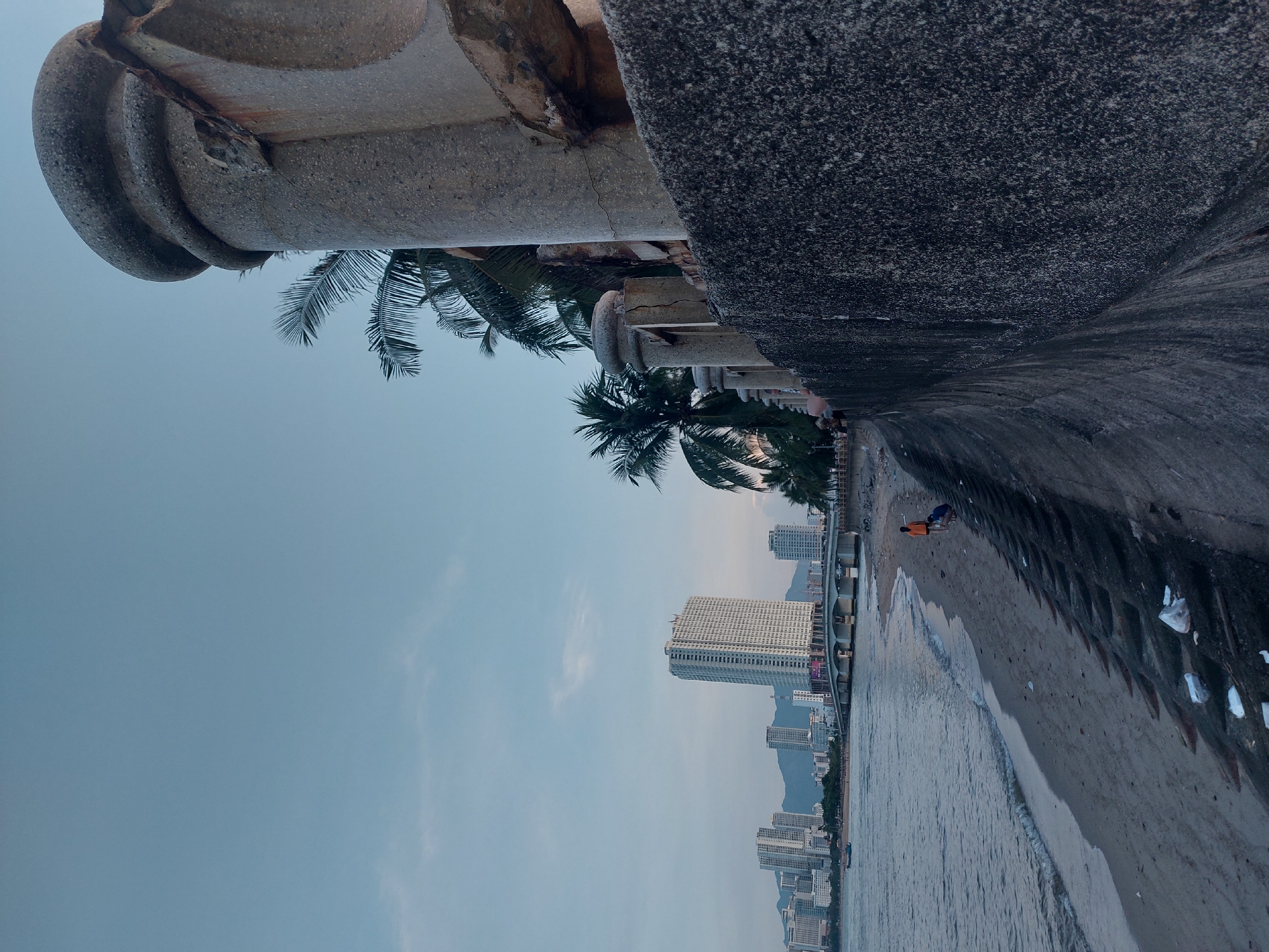 The image depicts a beachfront scene in Nha Trang, Vietnam, likely during dusk. In the foreground, a textured, grey concrete wall runs along the right side of the frame, with a stone pillar and railing overhead. Beyond the wall, a sandy beach stretches out, littered with plastic waste. The gentle waves of the sea lap the shore on the left. In the mid-ground, two individuals are present: one wearing an orange shirt and the other in blue shorts, appearing to interact on the sand. Further in the distance, a cityscape is visible, characterized by numerous high-rise buildings and a bridge spanning the water, silhouetted against a pale, cloudy sky. Palm trees are scattered along the coastline, and a small boat is on the water. The overall impression is one of a coastal urban environment.