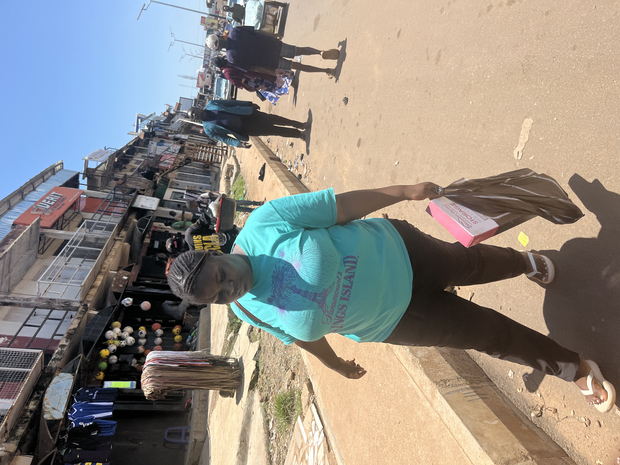 In a bustling street scene in Jos, Nigeria, a woman walks along a paved roadside lined with shops and stalls. She is wearing a teal t-shirt with a palm tree and Eiffel Tower graphic and dark trousers, and she carries a pink box and a folded brown bag. The street is moderately busy with pedestrians and vehicles, including cars and a cart being pushed. The shops on the left display various goods, including clothing and sporting equipment like soccer balls. The atmosphere suggests a typical day in a Nigerian urban market setting.