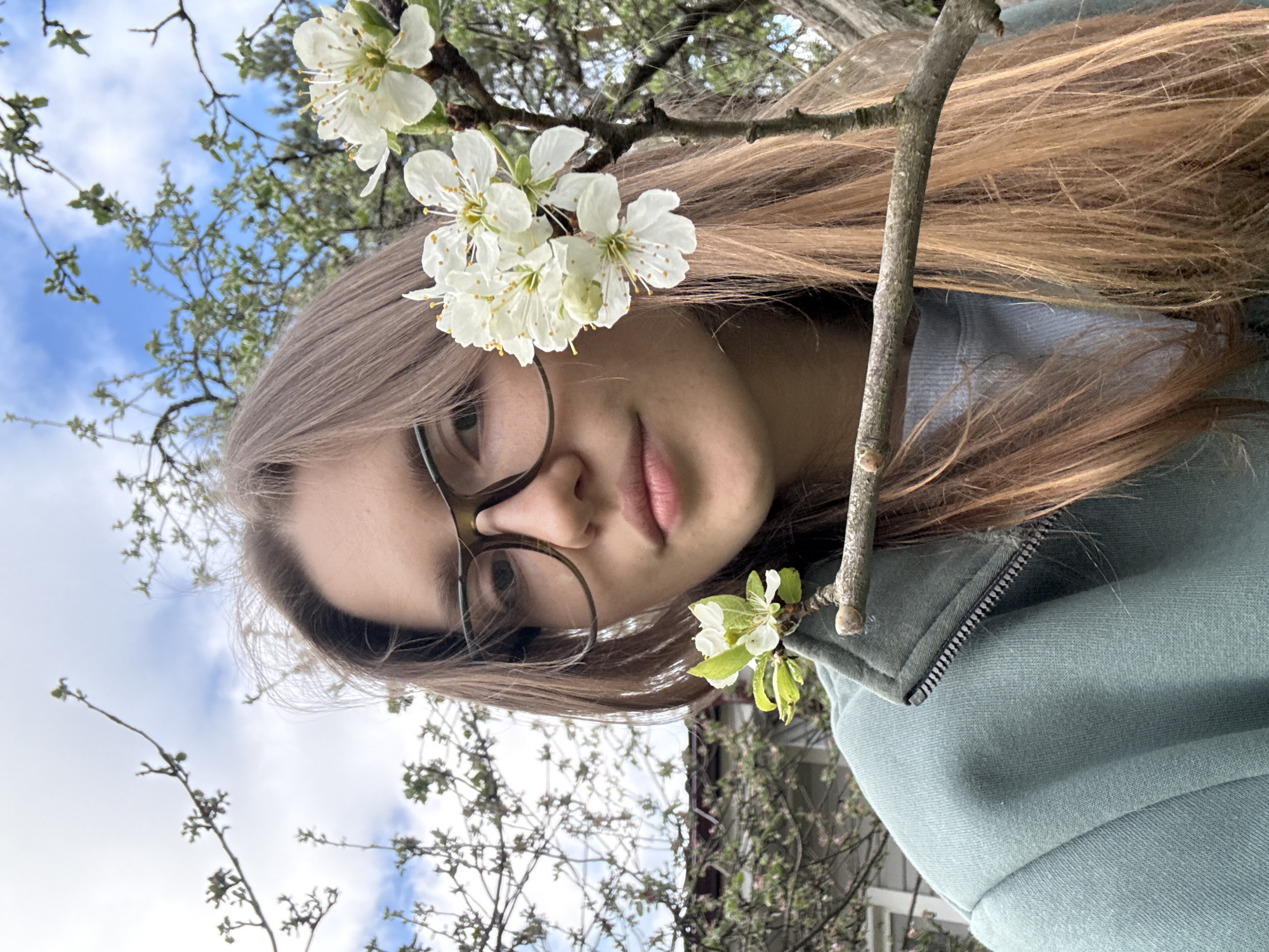 A young woman with light brown hair and glasses is in the foreground, partially obscured by a thin tree branch with white blossoms. The branch crosses in front of her face. She is wearing a light green or blue-gray hooded sweatshirt or jacket. The background shows more tree branches with sparse green leaves, a blue sky with some white clouds. The location, Ленинградская, Russia, cannot be confirmed with visual analysis.