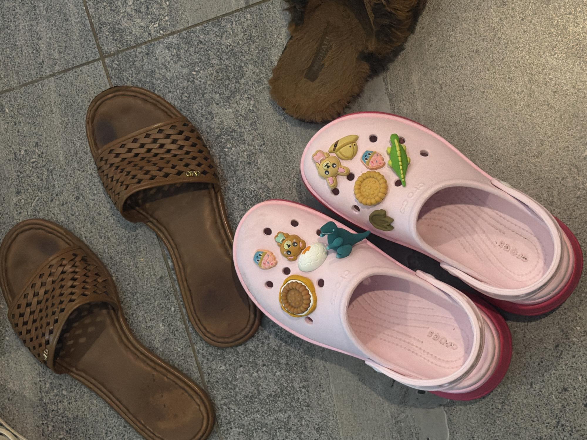 The image shows four pairs of shoes on a gray tile floor. Two pairs are brown woven-style sandals arranged side-by-side, one is a pair of brown fluffy slippers, and the other is two pink Crocs adorned with Jibbitz charms, including a dinosaur, cookie, and cupcake. The photo was taken in a space that is reminiscent of Kerobokan, Indonesia based on the type of flooring and surrounding details.