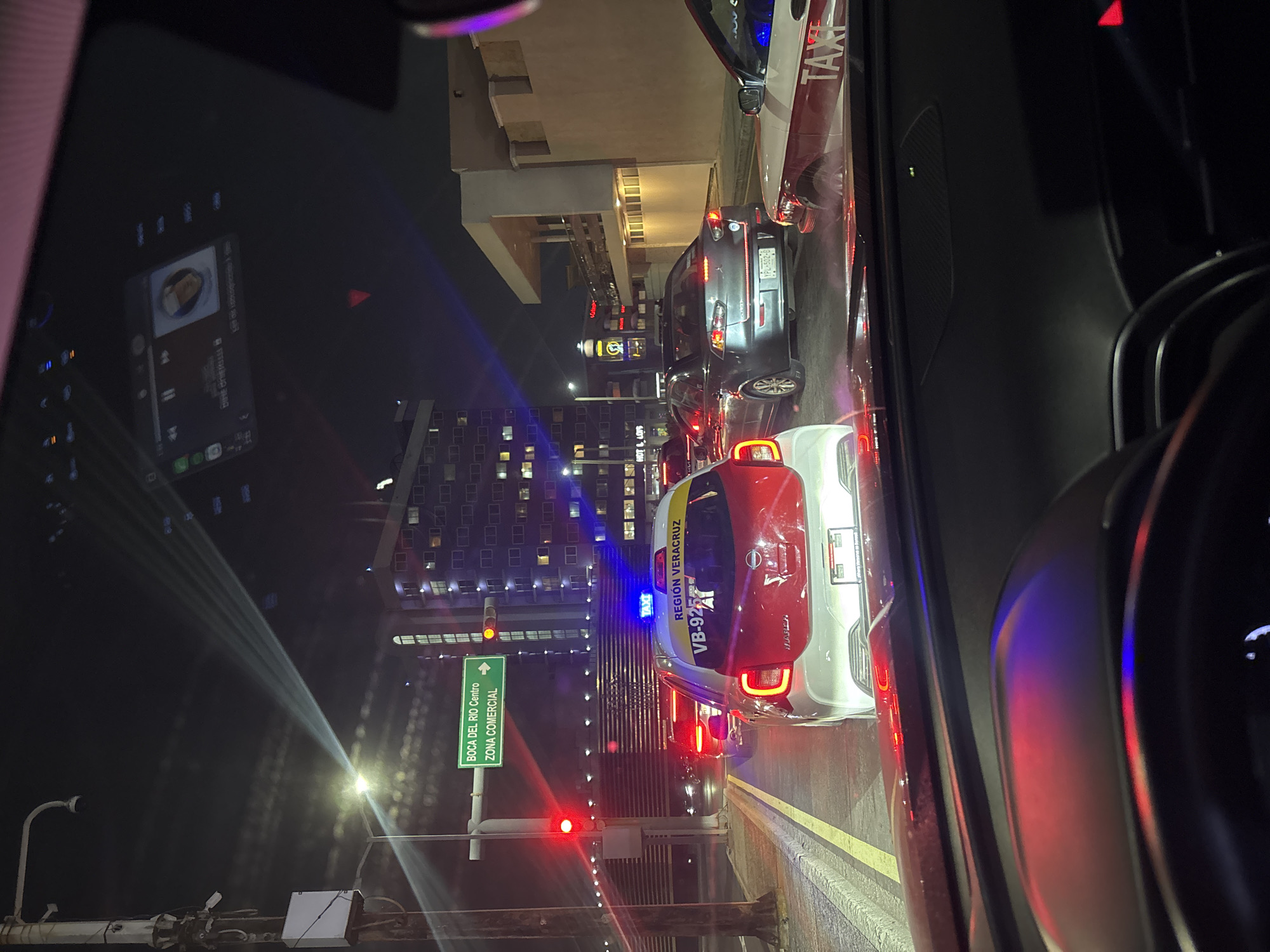 The image captures a nighttime urban scene from inside a vehicle, likely stopped in traffic. A prominent white and red SUV, possibly a police or patrol car, is positioned ahead with a blue light flashing on its roof. Visible text on its side reads "REGIÓN VERACRUZ" and "VB-92", along with a brand name that appears to be "MARU". Further down the street, other vehicles are visible, including a dark gray sedan and a taxi with "TAXI" printed on its side.
On the left side of the frame, a green traffic sign points towards "BOCA DEL RIO Centro" and "ZONA COMERCIAL", indicating a commercial district. A red traffic light is visible below the sign, confirming that traffic is halted. The background features several multi-story buildings with numerous lit windows, characteristic of a busy city at night. A sign for "HOT & LOIS" can be seen on one of the buildings.
A reflection of a smartphone screen displaying a music player interface is visible in the upper left portion of the car's windshield. The roads appear dry, and the night sky is dark, suggesting clear weather. The overall setting is a bustling street in an urban center, likely in Veracruz, Mexico, given the visible text cues. No people are distinctly visible.