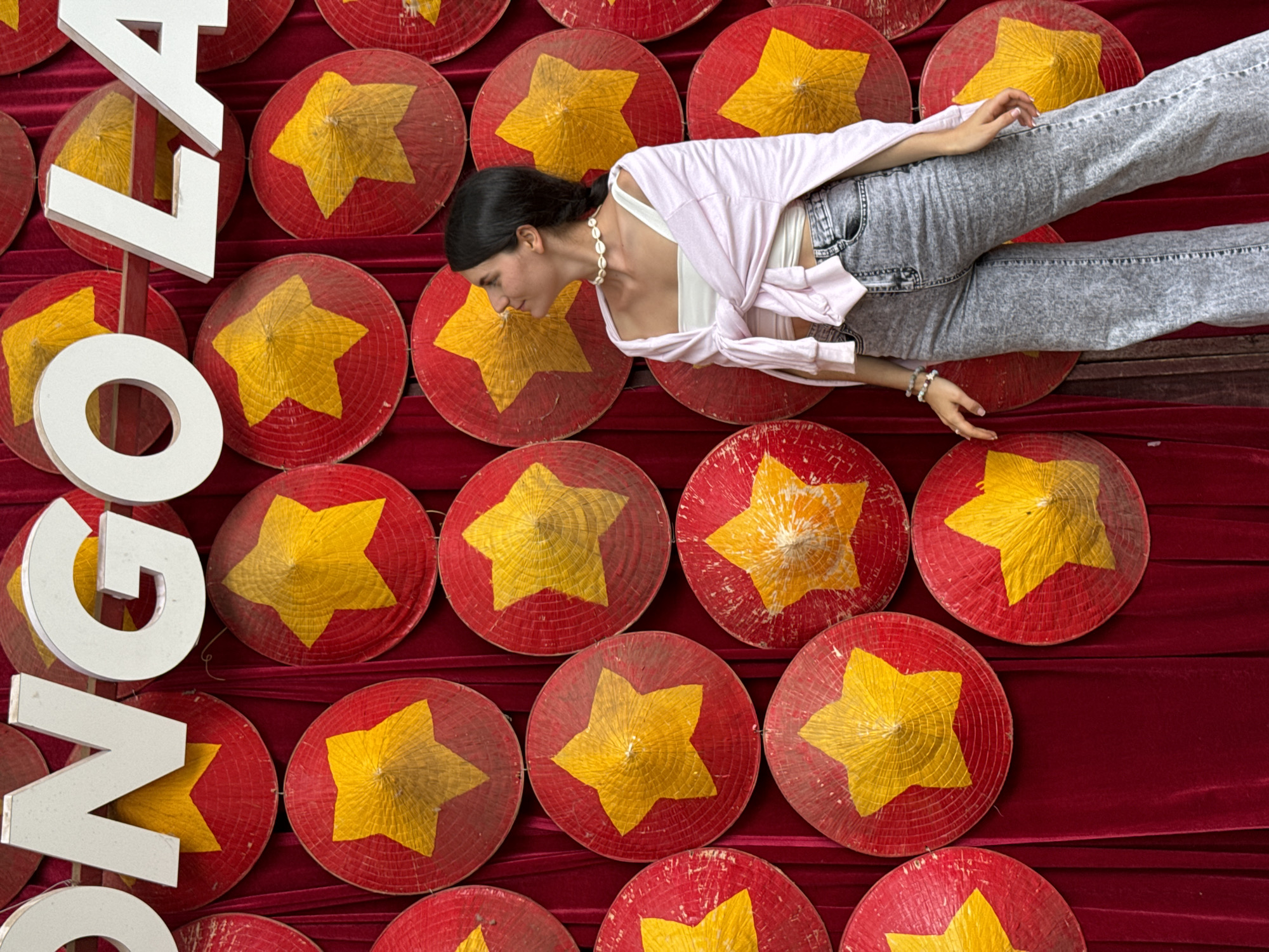 A high-angle, medium shot depicts a woman lying on a red velvet surface adorned with many red Vietnamese conical hats, each featuring a large, centrally placed yellow star. The woman wears a white tank top, a light pink, tied-front cardigan, and acid-wash jeans. She has dark hair pulled back in a ponytail and is wearing a shell necklace and a bracelet on her left wrist. Her body is angled across the scene, with her feet at the upper right corner and her head turned towards the left. Large, white, three-dimensional letters spelling out "NGOLA" are stacked to the left of the woman, partially obscuring the conical hats. The scene is located in Thôn 5, Vietnam.