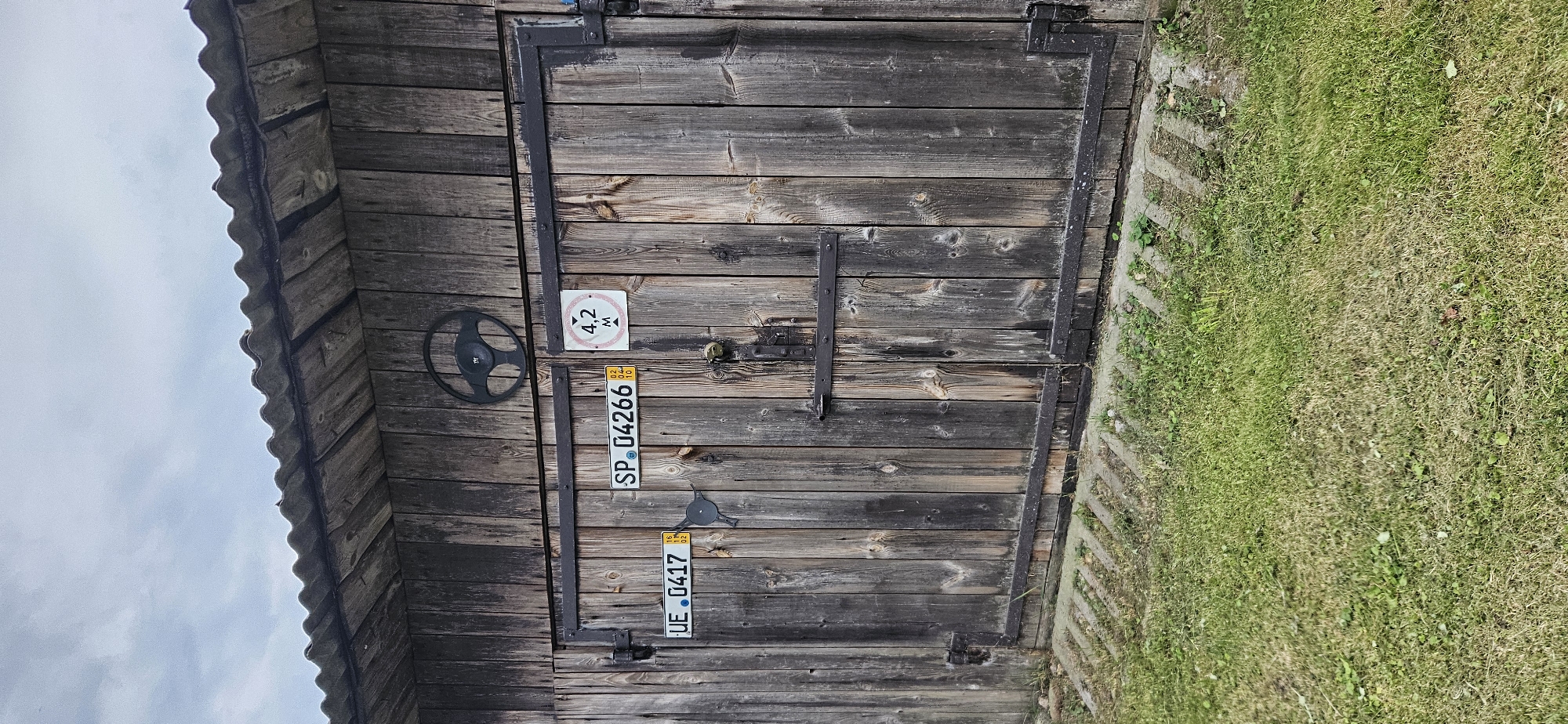 The image shows a weathered wooden shed with double doors, located in Мазалаўскі сельскі Савет, Belarus. A metal steering wheel ornament is affixed to the upper part of the shed's facade, centered above the doors. Two license plates are visible on the doors: one reads "UE 0417" and the other "SP 04266". To the right of the "SP 04266" plate, a square sign with "4,2 M" and an arrow pointing down is attached. The doors are secured by horizontal metal bands and a padlock. A small, dark bird-shaped object is mounted on the left door, below the "UE 0417" plate. The shed is situated in a grassy area with some concrete elements near its base. The sky is overcast. There are no people visible in the image.