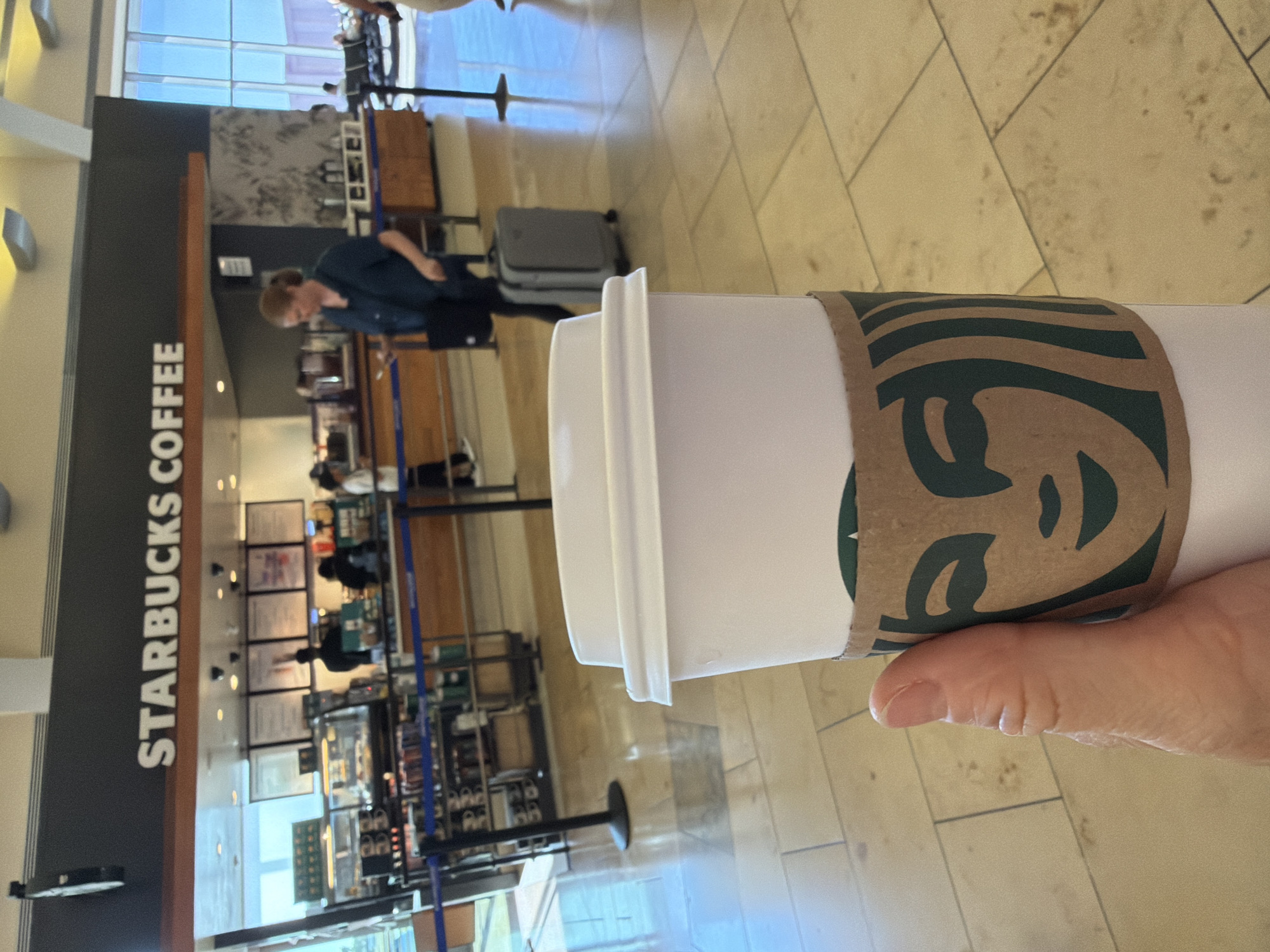 A close-up shot shows a hand holding a white Starbucks coffee cup with a brown sleeve featuring the Starbucks siren logo. In the background, a Starbucks coffee shop is visible, with people ordering and staff behind the counter. The location appears to be an airport terminal in Santa Ana, California, USA, indicated by the architecture and the presence of luggage. The environment is bright and modern, with tiled floors and large windows.