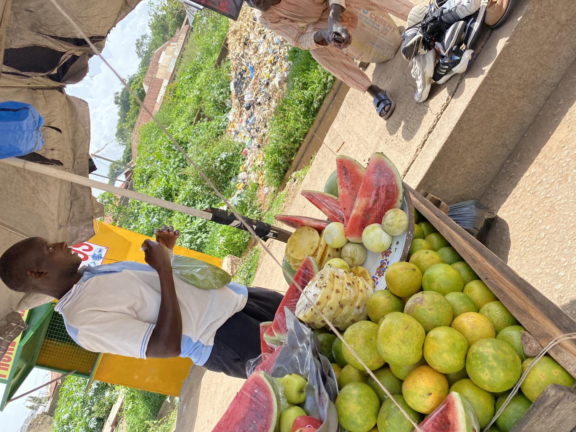 The image depicts an outdoor fruit stall located in Bukuru, Nigeria. A dark-skinned man wearing a blue and white collared shirt and dark shorts stands behind a display of fruits. He is holding a clear plastic bag containing green produce. The stall features a variety of fruits, including sliced watermelons, whole green apples, pineapples, and oranges. The fruits are arranged in wooden crates and on a metal tray. In the background, a pile of refuse and vegetation is visible, along with corrugated iron roofs of buildings on a hillside. To the right, another person, wearing a light-colored, patterned garment and dark sandals, sits near the stall. A stack of shoes is placed on the ground next to this person, and a large bag, possibly of cement, is also present. The stall is partially covered by a torn, off-white tarp, and a blue plastic object is suspended above. The ground is a sandy, reddish-brown color.