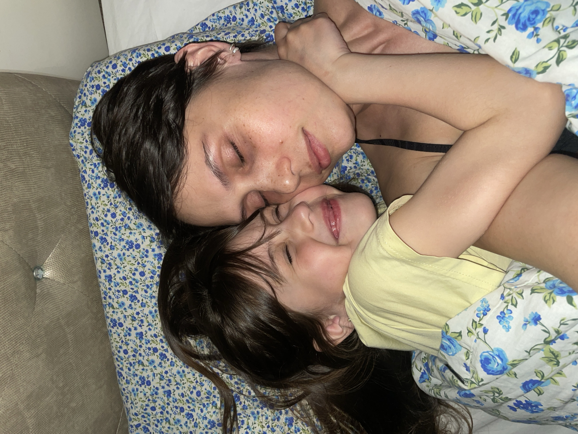 A woman and a young child are lying on a bed together, with their eyes closed. The woman is on top, with her face resting close to the child's. They are both under a blue and white floral patterned blanket. The woman has dark hair pulled back and is wearing a black top. The child has long brown hair and is wearing a yellow shirt. The headboard of the bed is visible in the background. The location cannot be determined from the image.