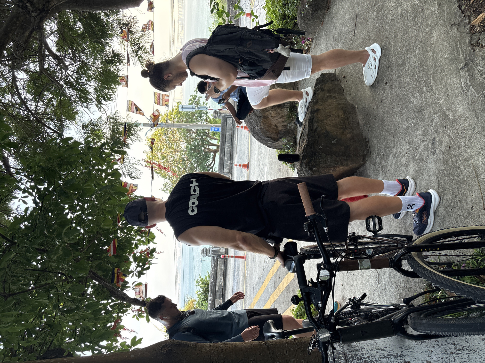 The image shows three people near the coast in what appears to be Đà Nẵng, Vietnam. In the foreground, a man wearing a black "COACH" shirt, black shorts, a cap, and white socks with blue DC shoes is positioned with a bicycle. Another man, wearing a long-sleeved grey shirt and black shorts is reclining against a tree trunk, potentially resting. A third person with a bun hairstyle is seen further away on a concrete surface, wearing a pink shirt, white shorts, white shoes, and carrying a black backpack, examining something on their hands. The background reveals a coastal view, with flags hanging on a line, presumably as decorations. The surroundings include trees, foliage, and rocky areas on the side.