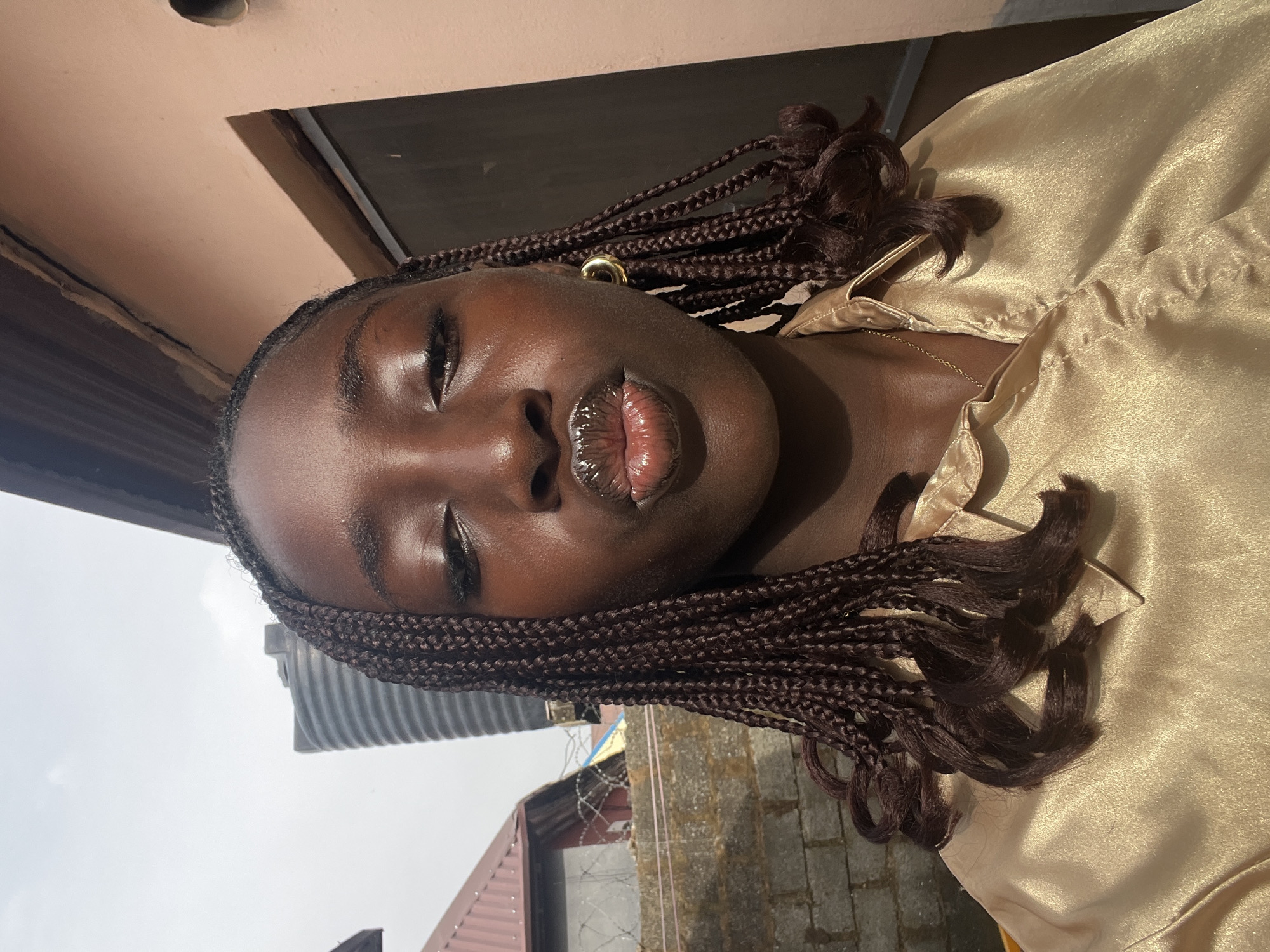 A close-up, upward-angle shot depicts a young Black woman with dark brown braided hair, closed eyes, and glossy lips. She is wearing gold hoop earrings and a gold-colored satin shirt. The background includes parts of a building exterior with a light peach-colored upper section, and a view of the sky. A corrugated metal water tank, a brick wall topped with barbed wire, and the edge of a red roof are also visible. The location appears to be outdoors. The specific location in Abuja, Nigeria, is not discernible from the image alone.