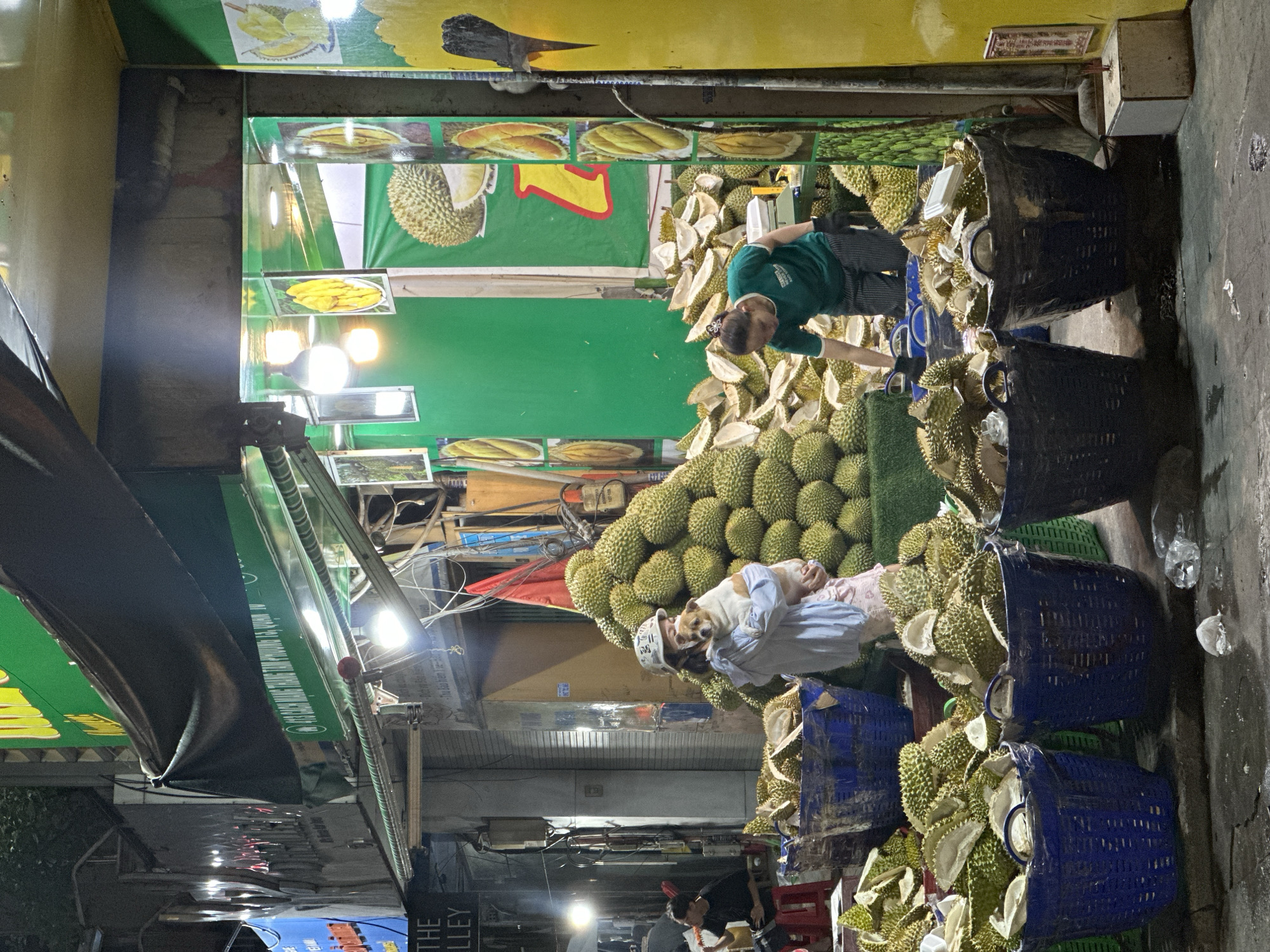 This image portrays a bustling durian fruit market located on a street in Thủ Đức, Vietnam. At the center of the scene, two vendors are selling durians; one is wearing a green shirt and examining durians, while the other, wearing a light-colored shirt, is interacting with a dog perched amidst a mound of durians. The market display consists of a large pile of whole durians and baskets overflowing with opened durians. The vendors are situated beneath a covered market stall, adorned with durian images. The street is narrow, bustling with people, and lined with various shops and buildings.