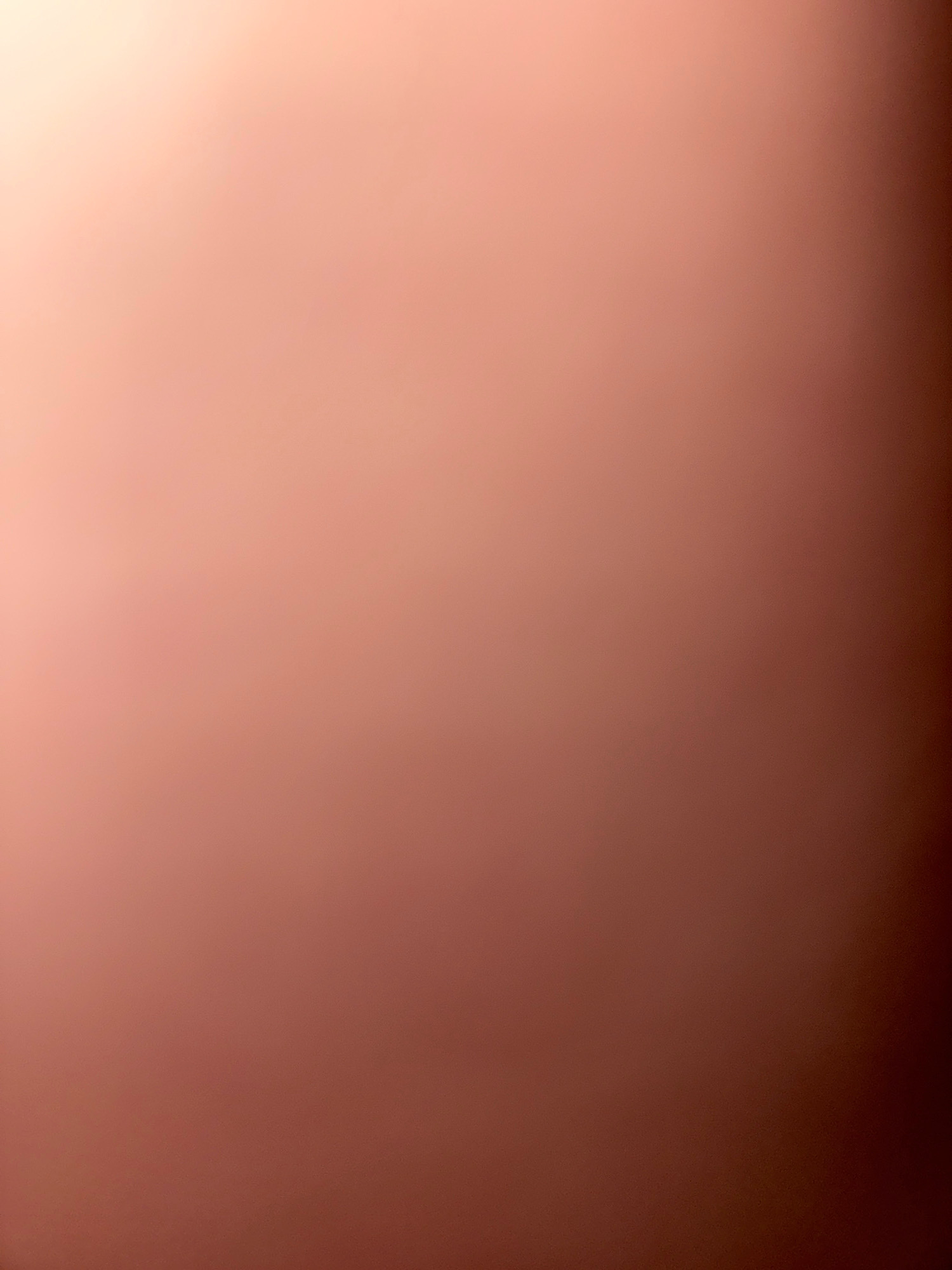 The image is an abstract, out-of-focus photograph of a surface with a gradient of colors. The background is predominantly a warm, peachy-pink hue that transitions to a deeper reddish-brown in the lower right corner. There are no discernible people, objects, actions, interactions, or environmental features visible in the image. It appears to be a close-up shot of a textured surface, possibly skin or fabric, illuminated by soft light that creates a luminous glow in the upper left quadrant. The lighting is diffused and creates subtle variations in tone across the surface. The image does not contain any identifiable elements related to the city of Zavolzhye, Russia.