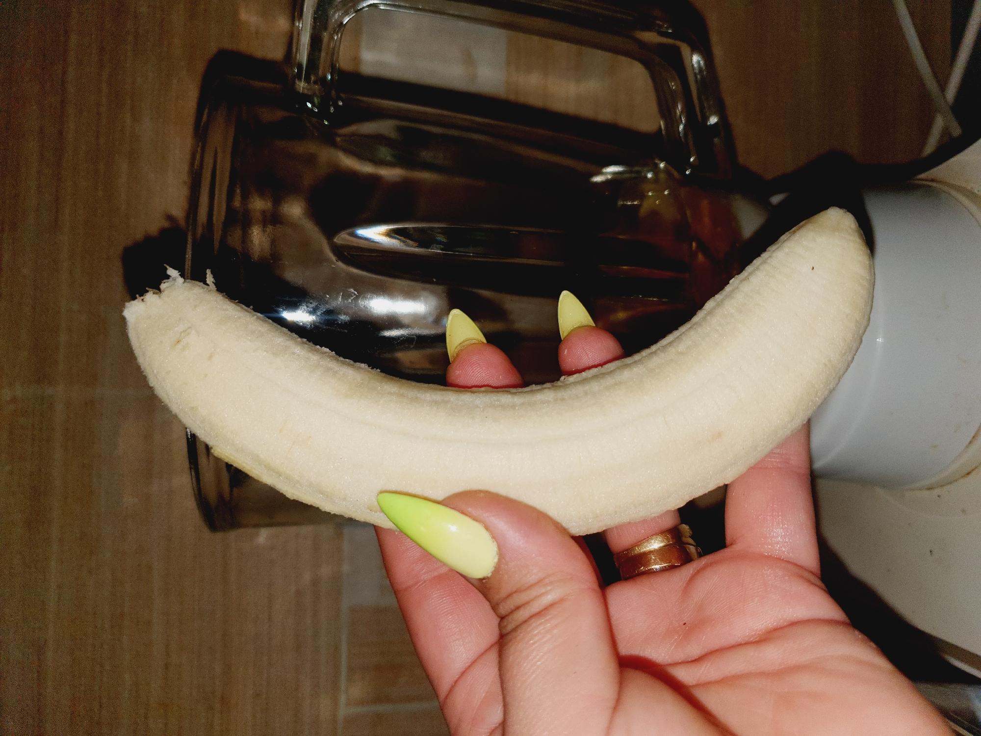 A close-up shot shows a person's hand holding a peeled banana over the opening of a blender. The banana is pale yellow and slightly curved, with a rough texture where it has been peeled. The hand has bright yellow, pointed fingernails with a gradient of green at the tips, and a gold ring is visible on the ring finger. The blender appears to be made of glass and plastic, with a dark liquid visible inside. The background consists of a tiled wall with a wood grain pattern. The scene is indoors, likely in a kitchen. There is no identifiable indication of the location being specifically in the city of Shiraz, Iran.