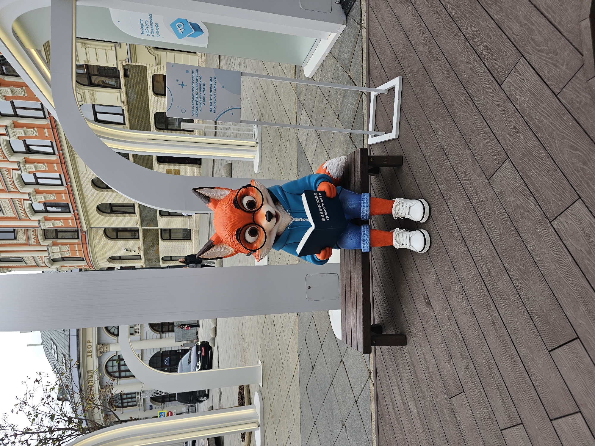 The image captures an outdoor scene in Moscow, Russia, likely in an urban public space. In the foreground, a cartoonish fox statue, dressed in a blue hoodie, denim shorts, and white sneakers, is seated on a wooden bench. The fox is holding an open book titled "Финансовая грамотность" (Financial Literacy). Behind the fox, there's a tall white modern structure with arched elements. To the right, a white stand displays informational text in Russian, with a blue logo featuring the number 10 and a stylized ruble symbol. In the background, a multi-story building with ornate architecture, featuring cream-colored walls and orange accents, stands prominently. Other elements visible include paved ground, trees with sparse autumn foliage, and a black car driving on a street. The overall setting suggests a well-maintained public area, possibly an exhibition space or a cultural hub in the city.