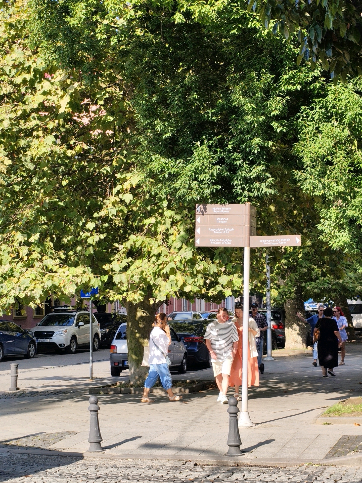 A street scene in Batumi, Georgia, features a signpost with directions to various museums, including the Adjara Museum, the Museum of Art, and the Archaeologist Museum. The signpost is situated near a large tree with lush green foliage, casting shadows on the paved sidewalk and street. Several cars are parked along the street, including a white SUV and a dark sedan. A group of people are walking on the sidewalk. In the foreground, a woman wearing blue pants and a white shirt is walking while talking on her phone. A man in a striped shirt and shorts is walking behind her, followed by a woman in a peach-colored dress. Further in the background, other individuals are seen walking and interacting. Buildings with pink facades are visible behind the parked cars. The overall atmosphere suggests a bright, sunny day in an urban environment.