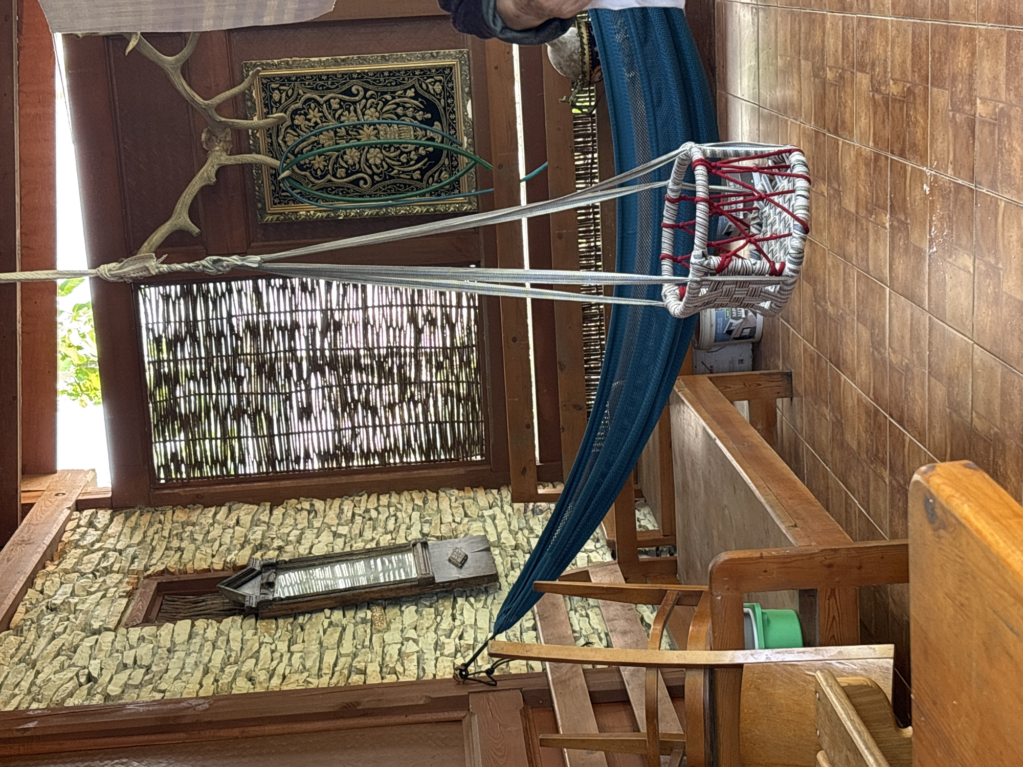 The image depicts a porch area in Gudauta, Abkhazia, featuring a blue hammock and a woven basket swing suspended from the ceiling. The porch has a tiled floor and walls constructed from light-colored stone. Wooden furniture, including benches and chairs, is visible. Decorative elements such as a framed tapestry and animal antlers are mounted on the wall. In the background, a woven window covering and a glimpse of foliage suggest an outdoor setting. The scene is illuminated by natural light.
