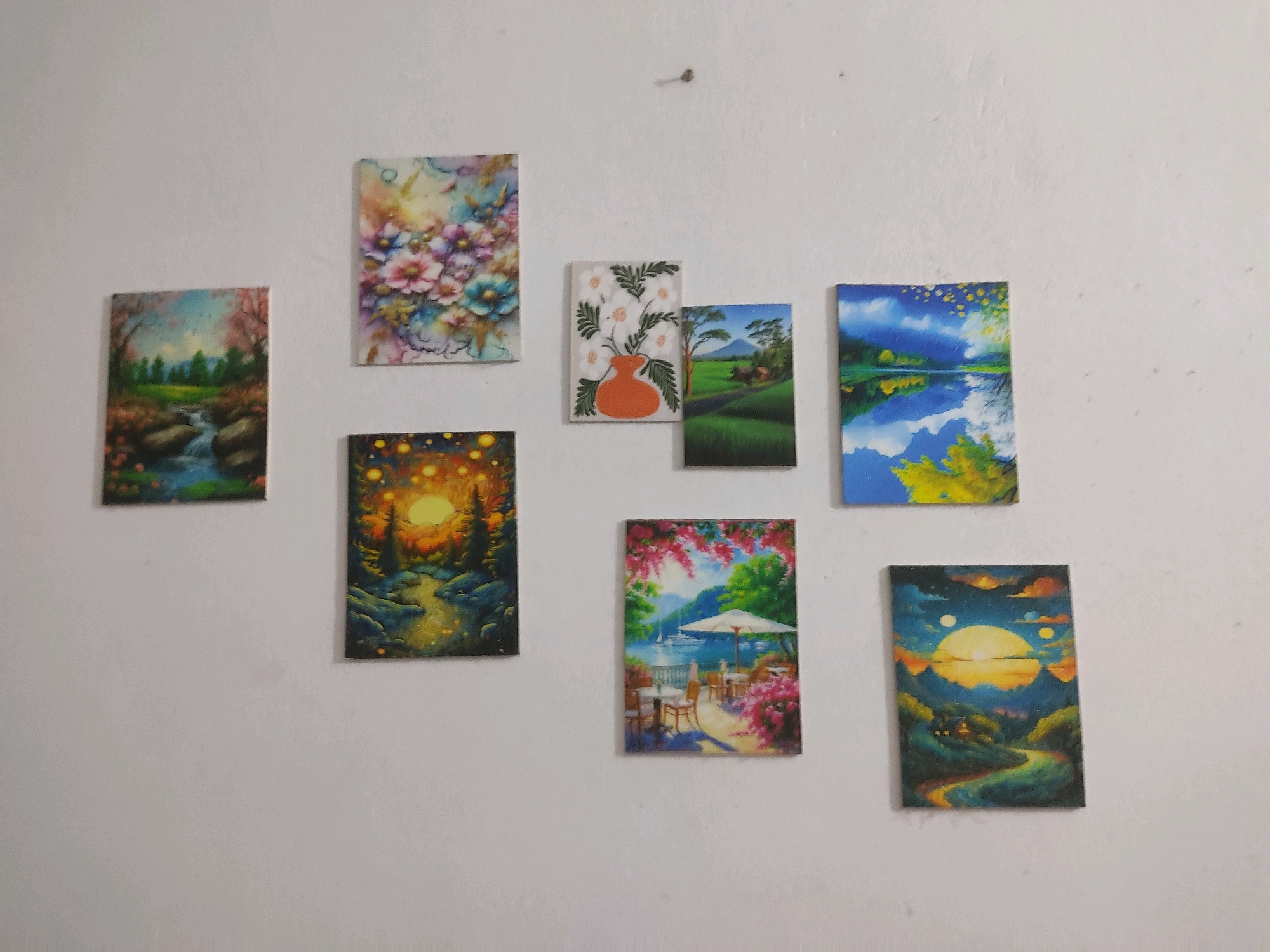The image shows a wall with eight small paintings of various landscapes and floral scenes. The paintings are arranged in two rows, with four paintings in the top row and four in the bottom row.
The top row paintings, from left to right, depict:
1. A floral arrangement with pink, blue, and yellow flowers against a textured background.
2. A vase with orange flowers and green leaves on a white background.
3. A green landscape with a body of water and a distant mountain under a blue sky.
4. A landscape with a body of water reflecting a blue sky with white clouds and yellow foliage.
The bottom row paintings, from left to right, depict:
1. A landscape with a stream flowing through a forest with trees and rocks, under a vibrant sky with a glowing sun.
2. A dark forest scene with a glowing sun or moon and a winding path.
3. An outdoor seating area with tables, chairs, and an umbrella, surrounded by pink flowers and lush greenery, with boats in the background on a body of water.
4. A landscape with a winding road, rolling hills, and a large sun with two smaller celestial bodies in a colorful sky.
The wall is white and appears to be textured. A small nail is visible in the upper center of the wall. The paintings are uniformly sized and appear to be framed or have a black border. The lighting suggests an indoor setting with light source from the front and slightly to the right.
There are no people, actions, or interactions visible in the image. The scene is a static display of artwork on a wall. The location is unspecified in the image itself, and no elements within the image indicate it is in Gazipur, Bangladesh.