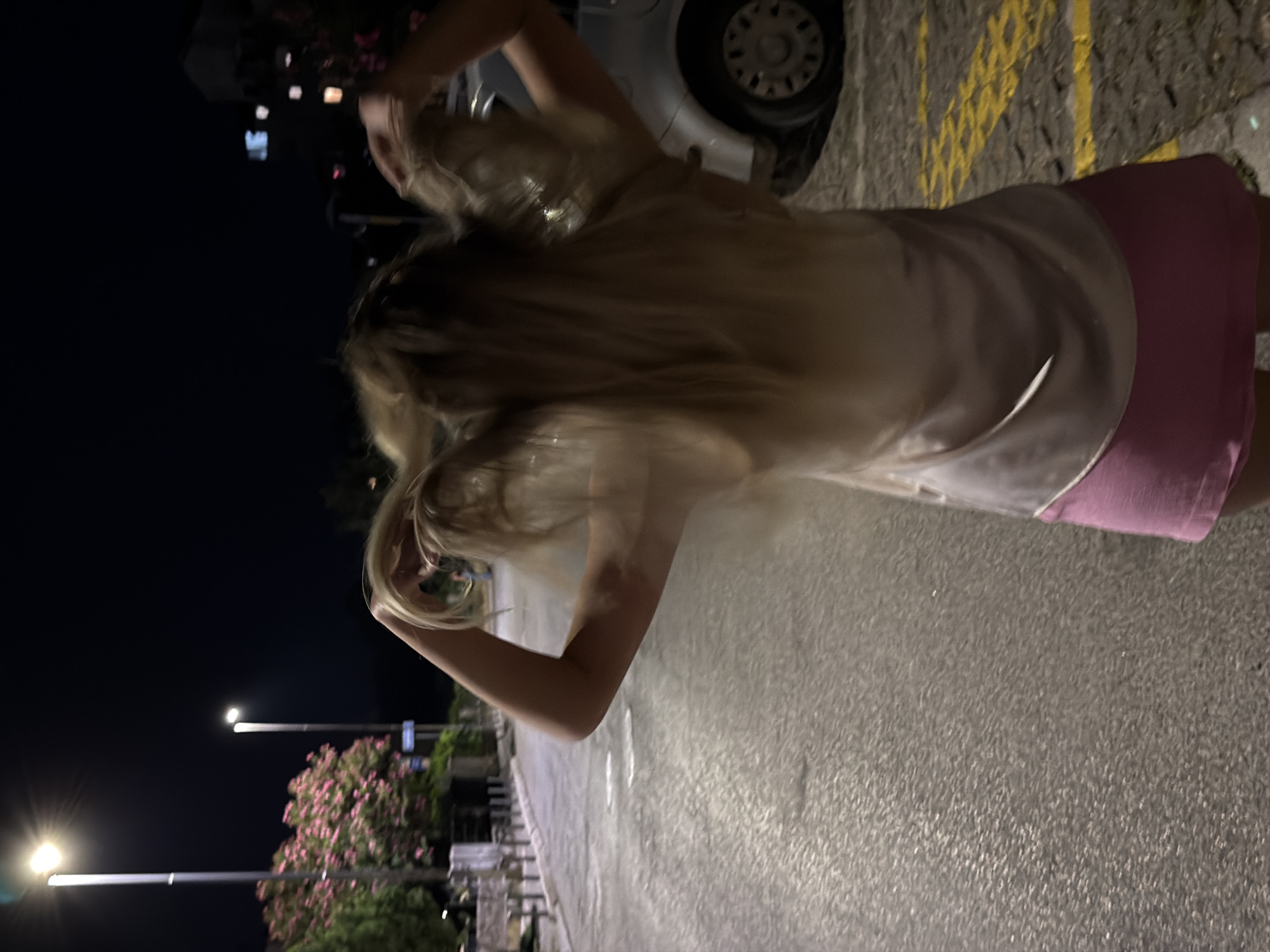 A medium shot captures a woman from behind, her blonde hair being tossed from side to side as she moves. She is wearing a light-colored top and a pink skirt. The setting appears to be a street in Budva, Montenegro, at night. A lamppost with a bright light illuminates the scene, and a tree with pink blossoms is visible on the left side of the image. A silver car is parked on the right, and yellow markings on the ground suggest a parking area or road delineation. The street is paved, and the overall ambiance is that of a warm evening in an urban environment.