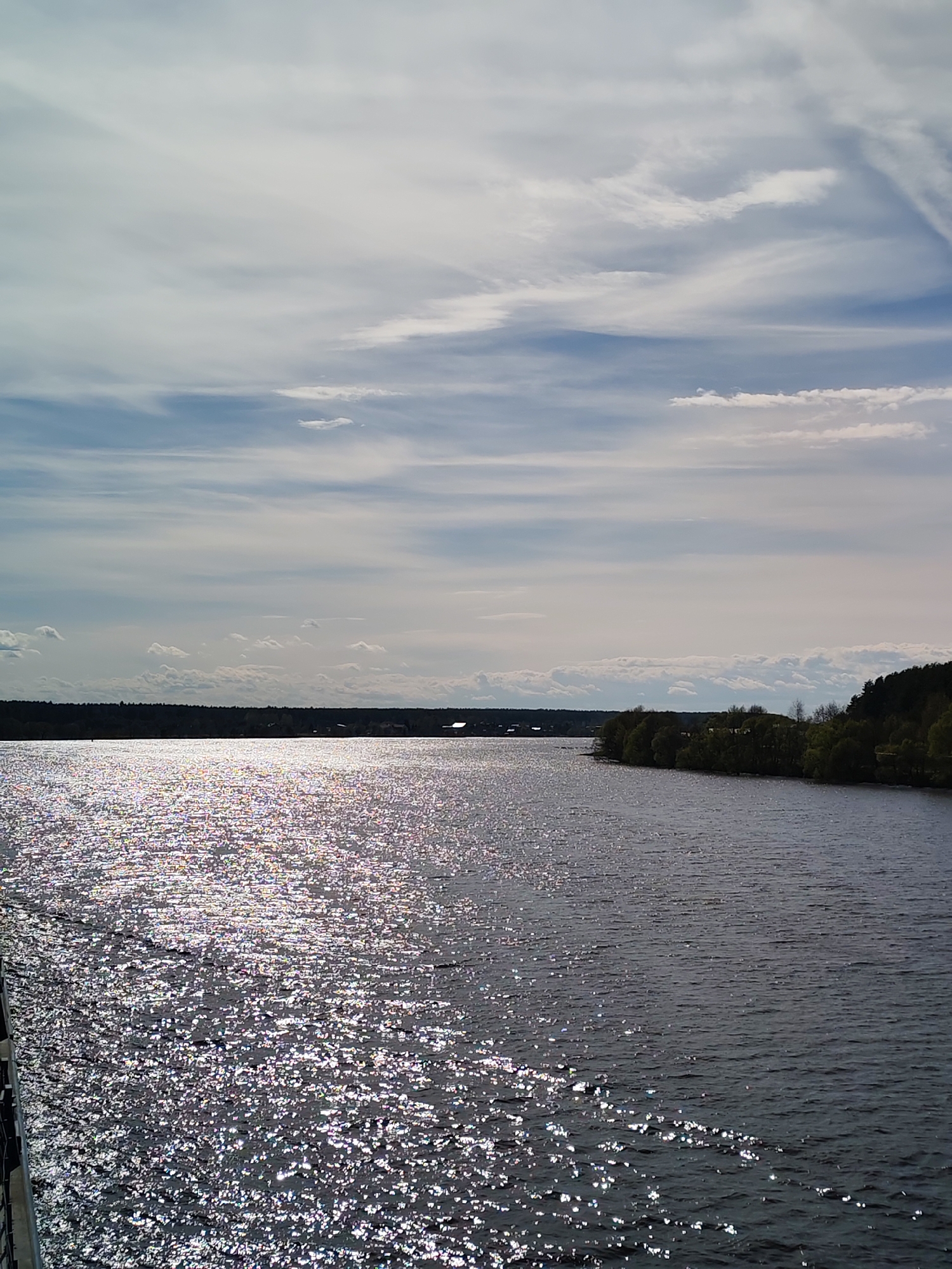 The image depicts a body of water under a sky with scattered clouds. Sunlight reflects off the surface of the water, creating a sparkling effect. The shoreline is visible in the distance, lined with trees and vegetation. The location is unspecified.