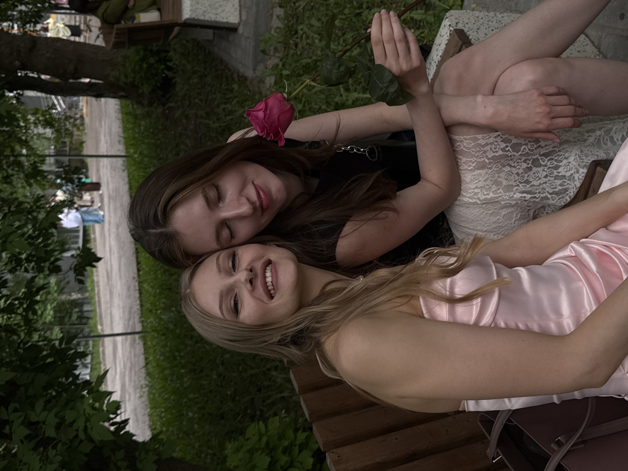 Two young women are sitting closely together on a wooden bench in an outdoor setting. The woman on the right has long blonde hair and is wearing a pink satin dress. She is smiling broadly, revealing her teeth. The woman on the left has long brown hair and is wearing a black top and a white lace skirt. She is holding a pink rose. The background shows green foliage, trees, and a walkway. Based on the style of architecture and the vegetation, this scene is likely located somewhere in the city of Perm, Russia.