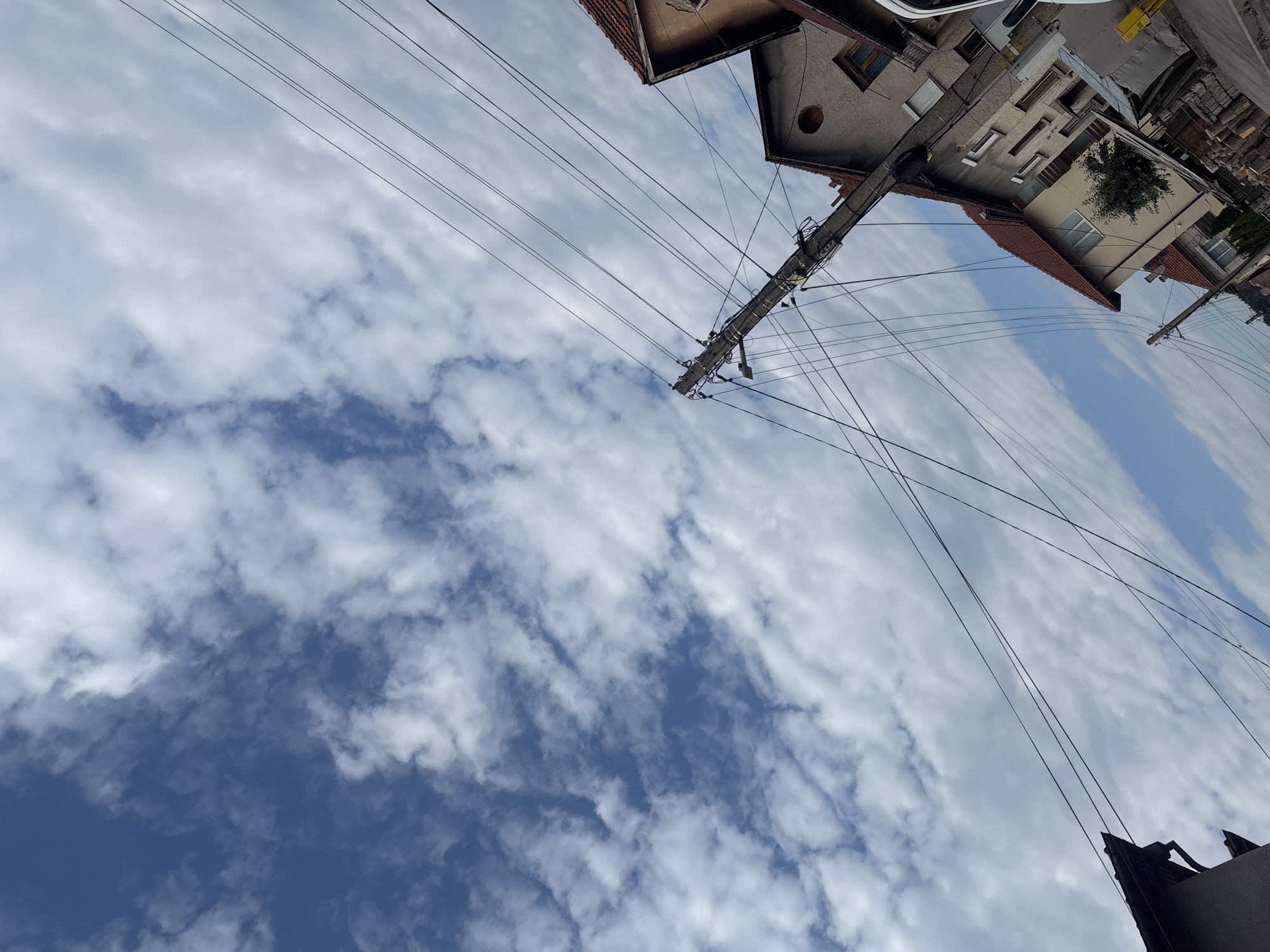 The image depicts an upward-facing view of a cloudy blue sky with utility poles and wires dominating the frame. In the upper right quadrant, parts of buildings are visible, featuring red-tiled roofs and cream-colored facades with windows. One building has a round, dark opening on its exterior. A prominent concrete utility pole, heavily laden with wires and smaller apparatus, is centrally positioned. The sky is filled with scattered white clouds against a deep blue background. The overall impression is of a residential or urban environment with overhead infrastructure. The lighting suggests it is daytime, possibly mid-morning or afternoon, with fair weather. The angle of the shot, looking straight up, emphasizes the sky and the complex network of wires. There is no visible text in the image. The location is identified as Samokov, Bulgaria.