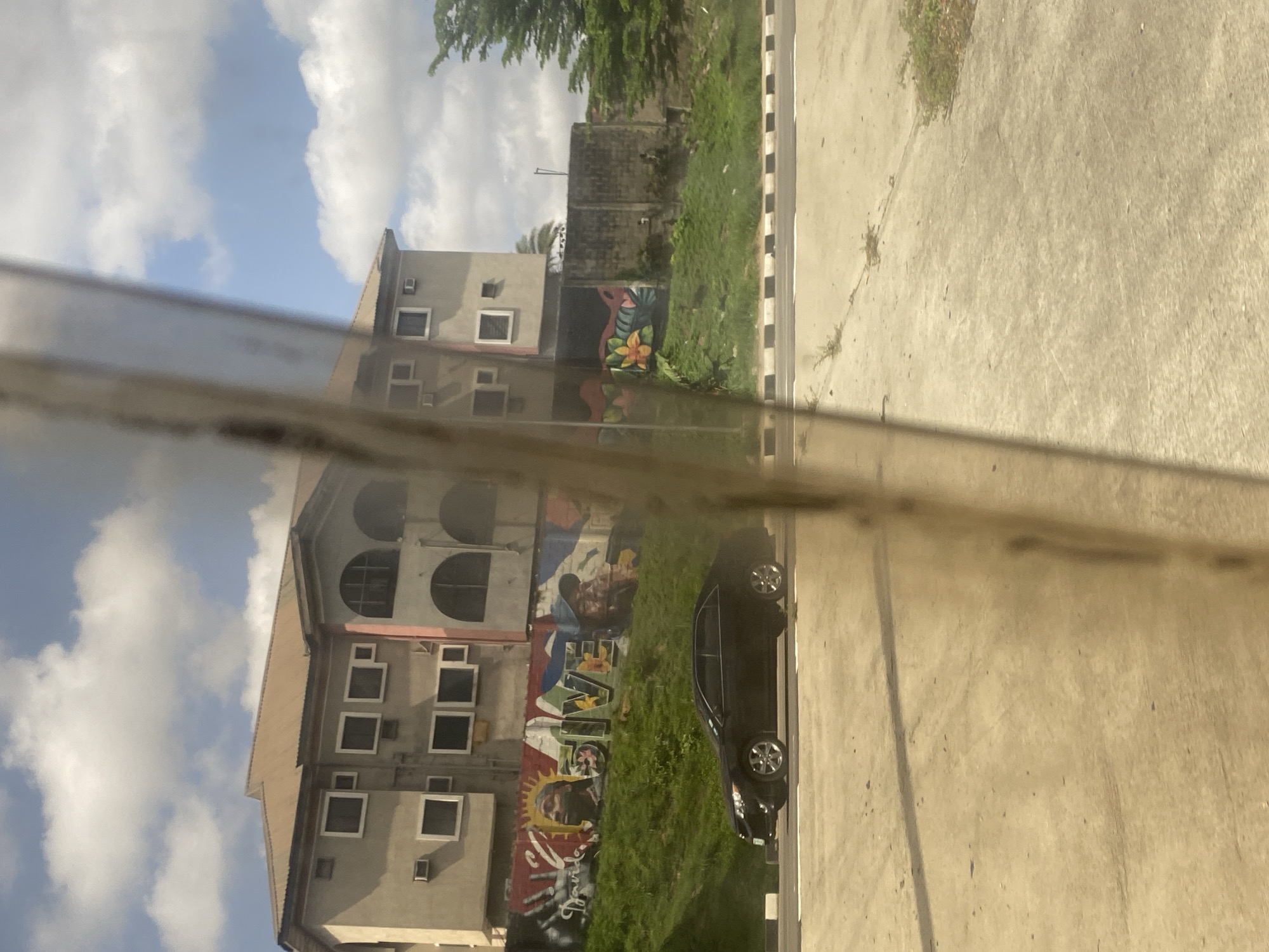 A slightly distorted, reflected image captures a multi-story building with several windows and a mural. Beside the building is a parked, dark-colored car on a paved surface near greenery. The sky above features scattered clouds. The location is Unknown, Unknown.
