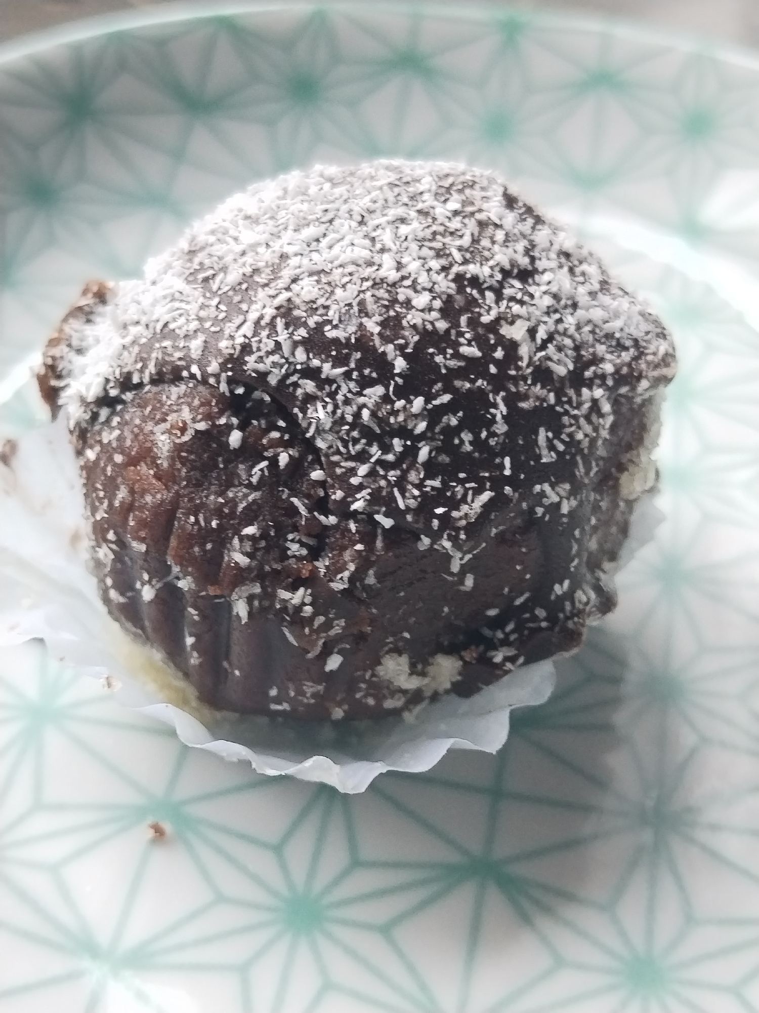 A single dark brown muffin or cupcake, covered in white shredded coconut flakes, is centered on a light-colored ceramic plate. The muffin is positioned within a white, fluted paper liner, which is slightly dislodged at the base, exposing a portion of the muffin's underside. The plate features a repeating geometric pattern of interconnected light teal or mint green star-like shapes against a white background. Small brown crumbs are visible on the plate near the base of the paper liner. The scene is illuminated by ambient light, creating soft highlights on the coconut and subtle shadows on the plate. This depiction takes place in Бараўлянскі сельскі Савет, Belarus.