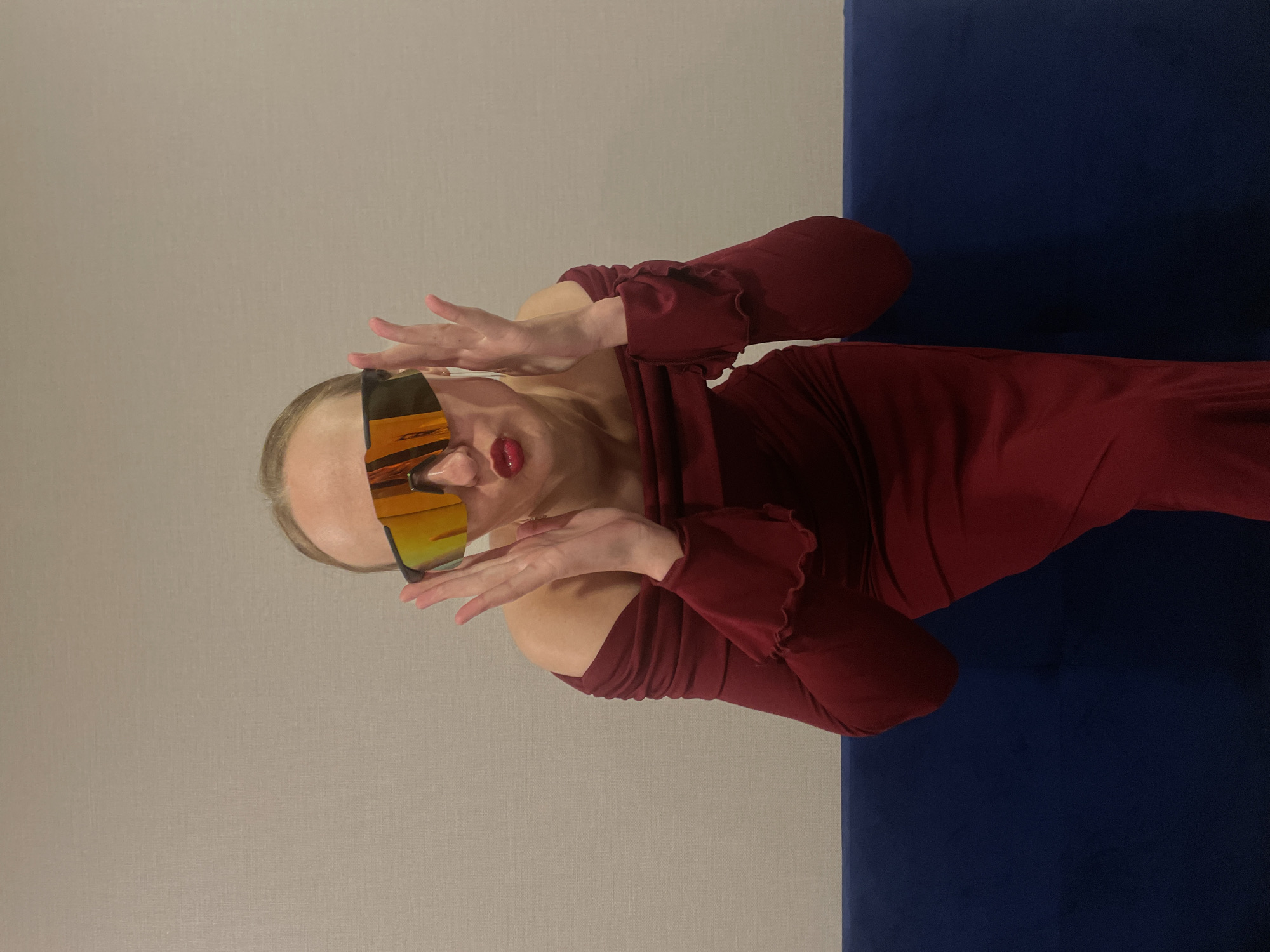 A woman wearing a maroon dress and futuristic gold visor sunglasses is positioned against a background consisting of a light grey wall on the left and a dark blue surface on the right. She is posing with her hands raised and curved near her face, seemingly surprised or making a playful gesture. Her red lipstick is a stark contrast to her fair skin. The image is slightly tilted. There is no information available to link this photo to Подшивалово, Russia.