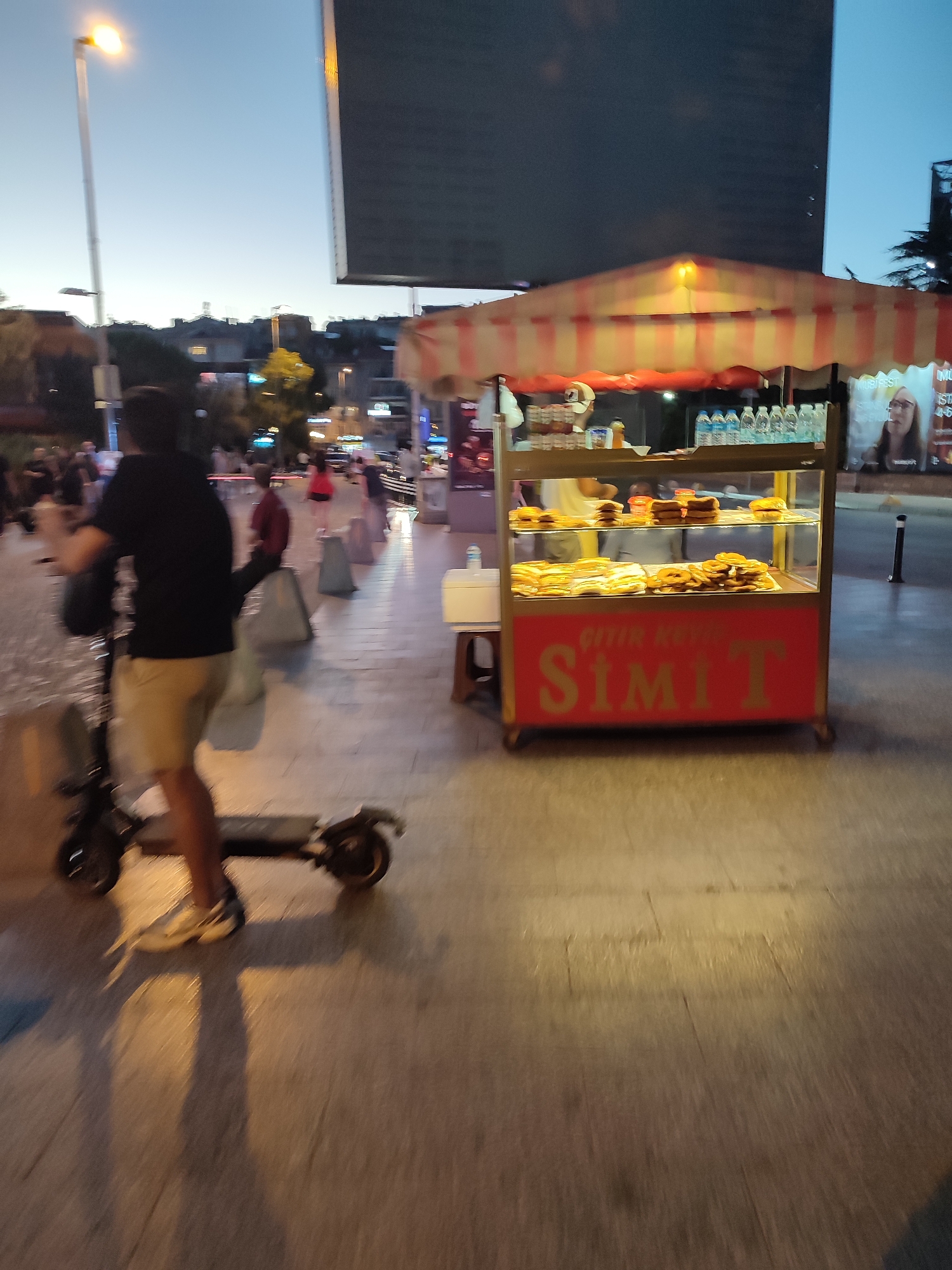 The image captures a street scene in Kadıköy, Turkey, at dusk. In the foreground, a person is riding an electric scooter from left to right, with their back to the camera. They are wearing a black t-shirt and tan shorts, and athletic shoes. The scooter is dark in color. Behind the scooter rider, a street vendor's cart is visible, displaying various food items, likely simit, a type of Turkish bread, as indicated by the prominent "SİMİT" text on the cart. The cart has a striped awning in red and yellow. To the right of the cart, a poster featuring a woman is displayed. The background shows other people walking along the sidewalk, some buildings, streetlights, and trees, all blurred due to motion and low light conditions. The overall atmosphere suggests a bustling urban environment transitioning into evening.