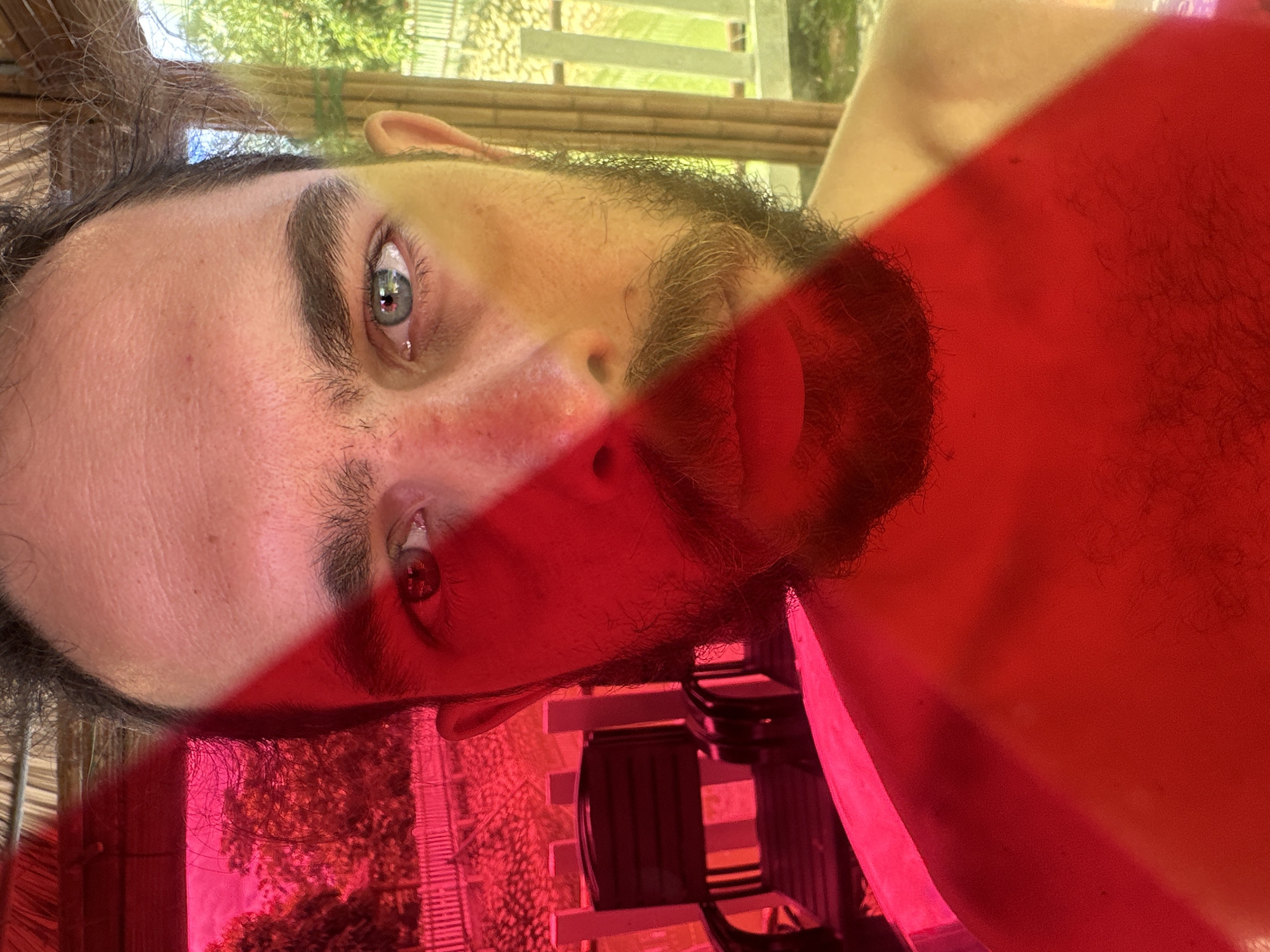 A male subject, with a beard and mustache, is presented in a close-up, chest-up portrait. A strong, diagonal beam of intense red light bisects the frame, covering the left half of the subject's face and body, and the background on that side. His left eye is obscured by the red light, while his right eye is clearly visible, blue/grey in color, with a distinct reflection. The right side of his face and bare chest are illuminated by natural light, showing a fair complexion. In the red-illuminated background on the left, an outdoor setting is discernible, featuring trees or dense foliage, a light-colored fence, and dark outdoor chairs. Above, a structure with a thatched or natural-material roof is partially visible. In the naturally lit background on the right, lush green foliage and light-colored vertical posts or fencing elements indicate an outdoor environment. The scene is situated in Nghia Lo Ward, Vietnam.