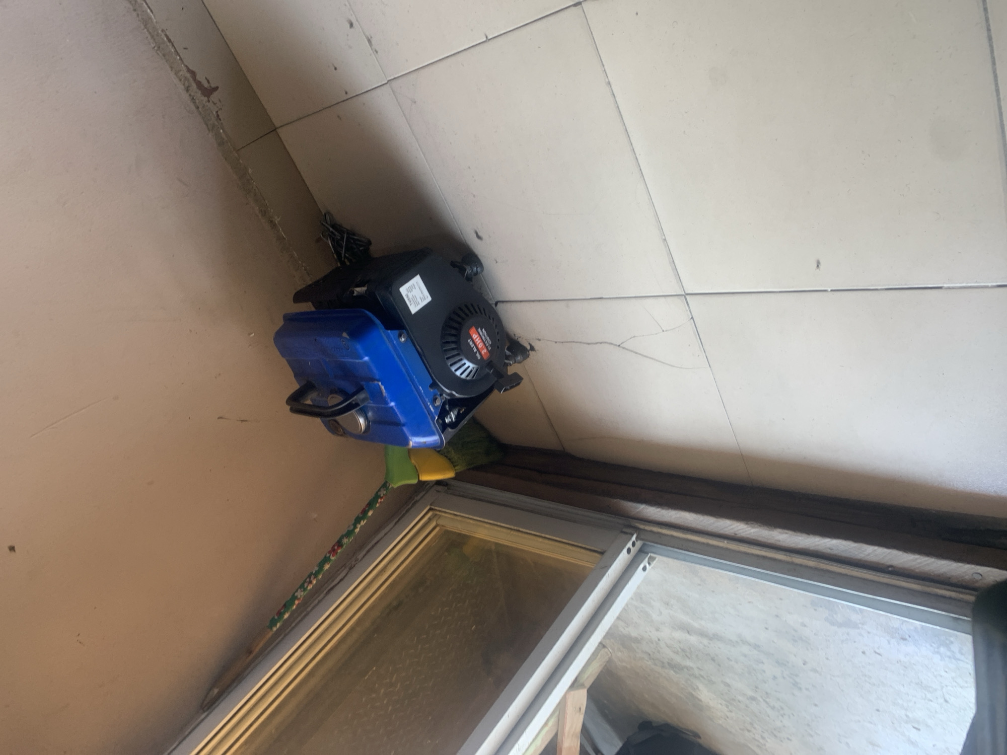In Jalingo, Nigeria, a blue and black portable generator is positioned on a tiled floor next to a doorway. The generator is small, with a black engine casing and a blue fuel tank. A broom with a green and patterned handle leans against the wall near the generator, its bristles touching the ground. The doorway appears to have a sliding glass panel and a wooden frame. The walls are painted a neutral color, and the floor is covered in light-colored square tiles. The overall scene suggests a storage area or a corner of a room within a building.