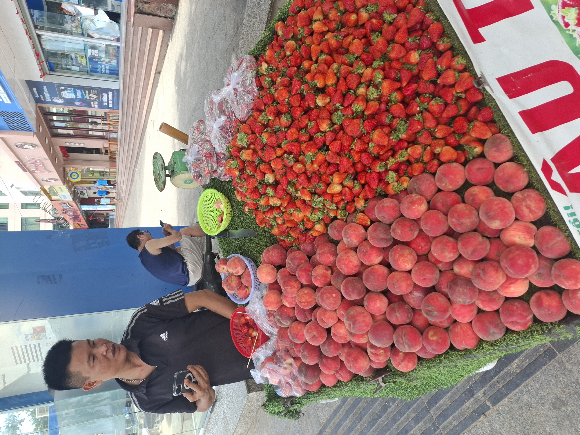 A Vietnamese man stands by a fruit stand displaying a large pile of peaches and strawberries. He is wearing a black polo shirt and holding a smartphone. The fruit stand is set up on a sidewalk in front of several shops. In the background, a second man is sitting and looking at his phone. The street appears to be in Nha Trang, Vietnam, as indicated by the signage on the buildings. The atmosphere is bustling, suggesting a commercial area.