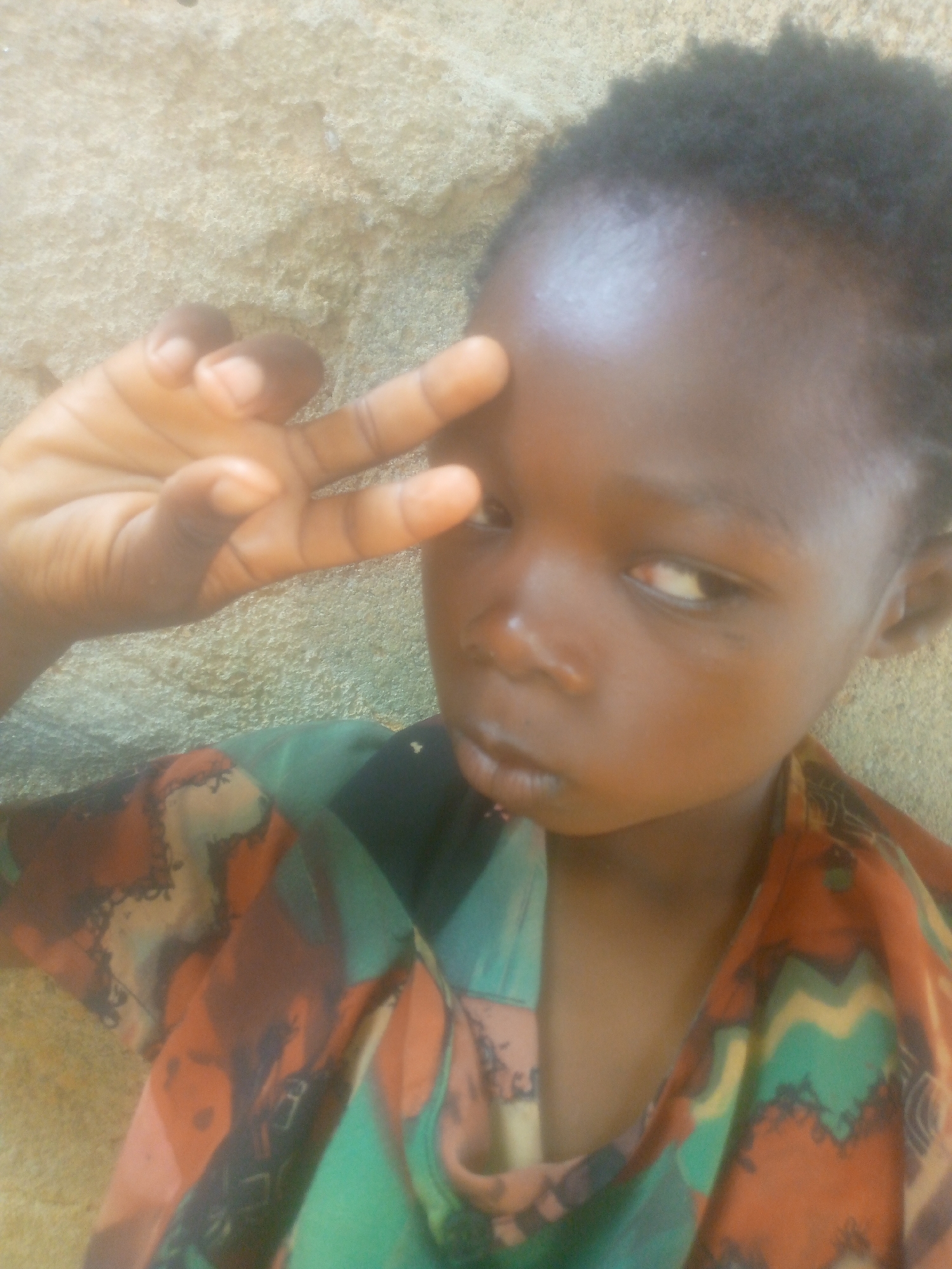 A young dark-skinned person poses for the camera, holding up two fingers in a peace sign near their forehead. They have short, dark hair and are wearing a patterned shirt with colors including green, orange, and brown. The background appears to be a textured wall. The city and region are unknown.