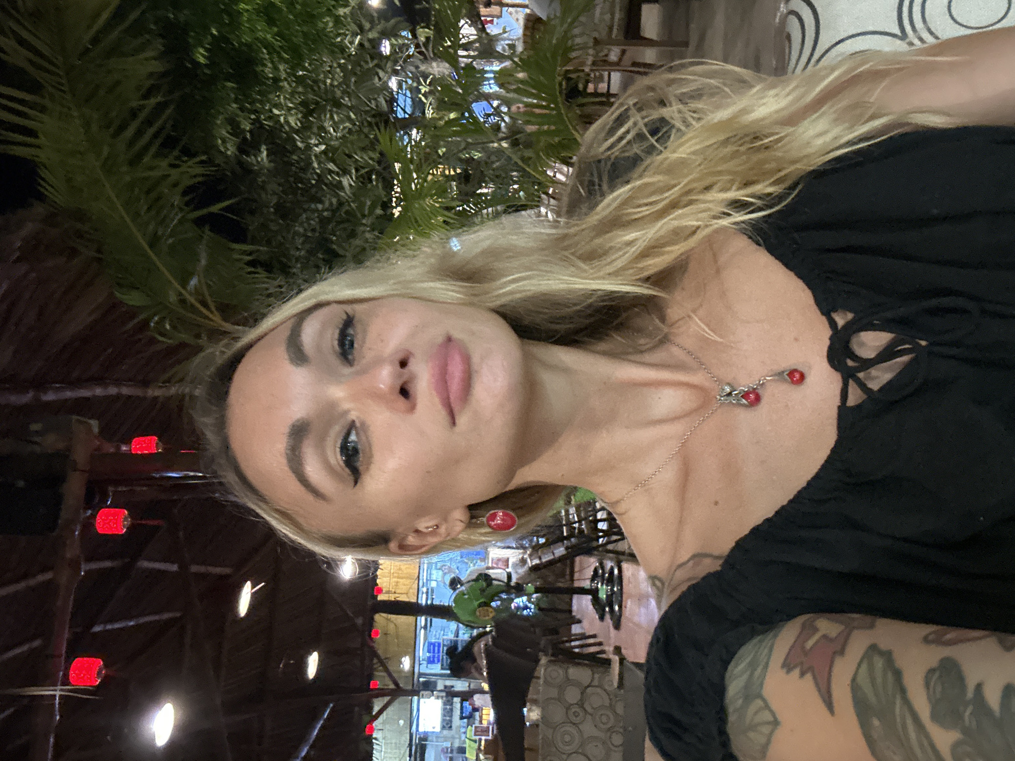 The image displays a selfie-style portrait of a woman in an evening setting. The main subject is a fair-skinned woman with long, wavy blonde hair, looking directly at the camera. She has noticeable dark eyeliner, full lips, and a neutral to slightly pouting expression. She is wearing a black, off-the-shoulder or peasant-style top. A delicate silver necklace with two red, cherry-like charms adorns her neck. Her left arm, visible in the foreground, is covered with various tattoos.
The setting is an outdoor or semi-outdoor restaurant or bar at night. Lush green palm trees are prominent in the upper and left background, some illuminated by artificial light. The venue features dark wooden structural elements, including beams and a ceiling made of natural materials. The background is dark, indicating nighttime, and is lit by numerous artificial lights; these include glowing red cylindrical or lantern-like fixtures and diffused white circular lights, creating a warm, inviting atmosphere. In the blurred mid-ground, hints of a bustling establishment are visible, including chairs, tables, and a bright green scooter or motorbike. A patterned gray and white upholstered surface is also partially seen.
The activity is a casual pose for a photograph, likely a selfie. The time of day is clearly night. No specific weather conditions are discernible, but the open-air setting suggests a warm climate. The location cues, such as palm trees, wooden structures, and lantern-style lights, are consistent with a tropical or Southeast Asian ambiance, aligning with the provided context of Phu Thuy Ward, Vietnam. There is no visible text in the image.