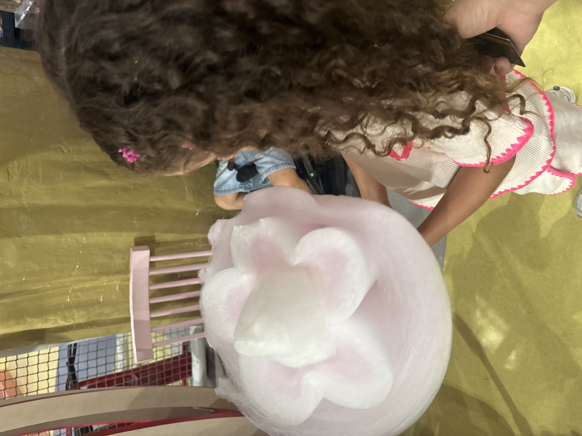 A young girl with curly brown hair, adorned with a small pink clip, is holding a large, fluffy pink and white cotton candy that is shaped like a flower. She is wearing a cream-colored top with bright pink trim. Her left arm is holding the cotton candy, and her right hand is holding a dark object, possibly a phone. The girl is facing away from the camera, looking towards a yellow wall in the background. To the left of the girl, a pink chair with a slatted back is visible. The floor is a bright yellow-green color. The overall impression is a candid moment of a child enjoying a treat in what appears to be an indoor entertainment or play area. The setting does not provide specific visual cues to pinpoint a location within Dubai, United Arab Emirates, but it suggests a recreational space.