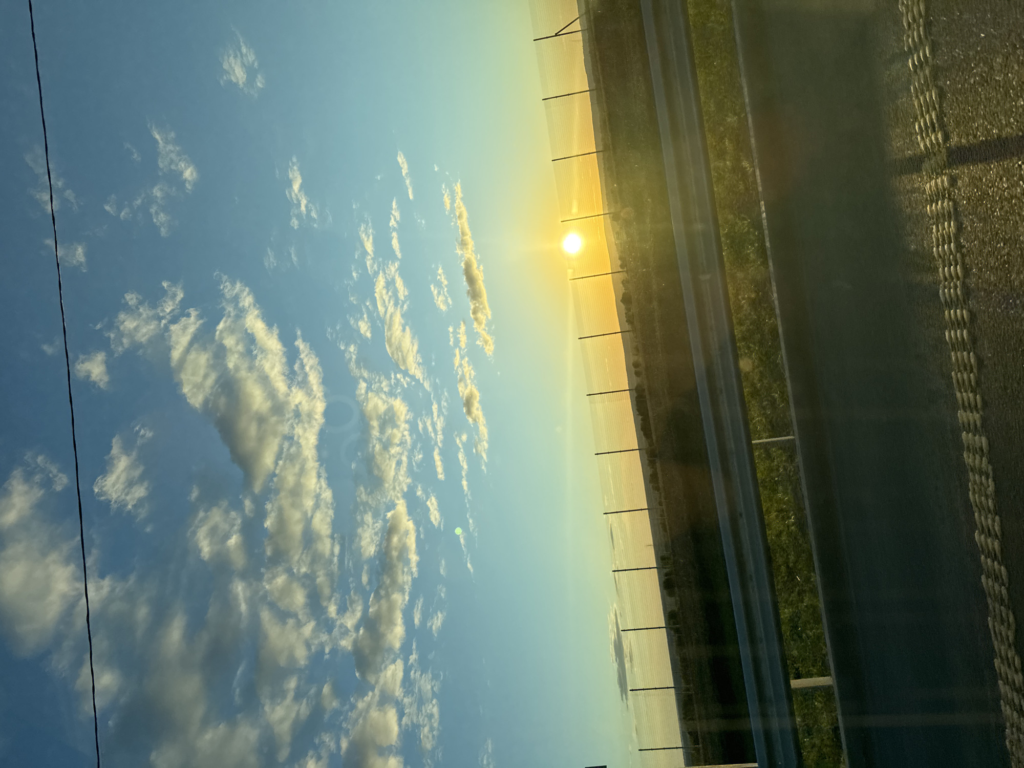 The image depicts a view from a vehicle, likely a car, moving along a highway at dusk or dawn, with the sun positioned on the horizon. To the left, the sky is filled with white cumulus clouds against a light blue sky. A power line is visible in the corner. To the right, a highway stretches out. A fence runs along the highway. The roadside is grassy. The road surface is dark. The specific location in Kazy-El'dyak, Russia, cannot be confirmed without additional contextual information.