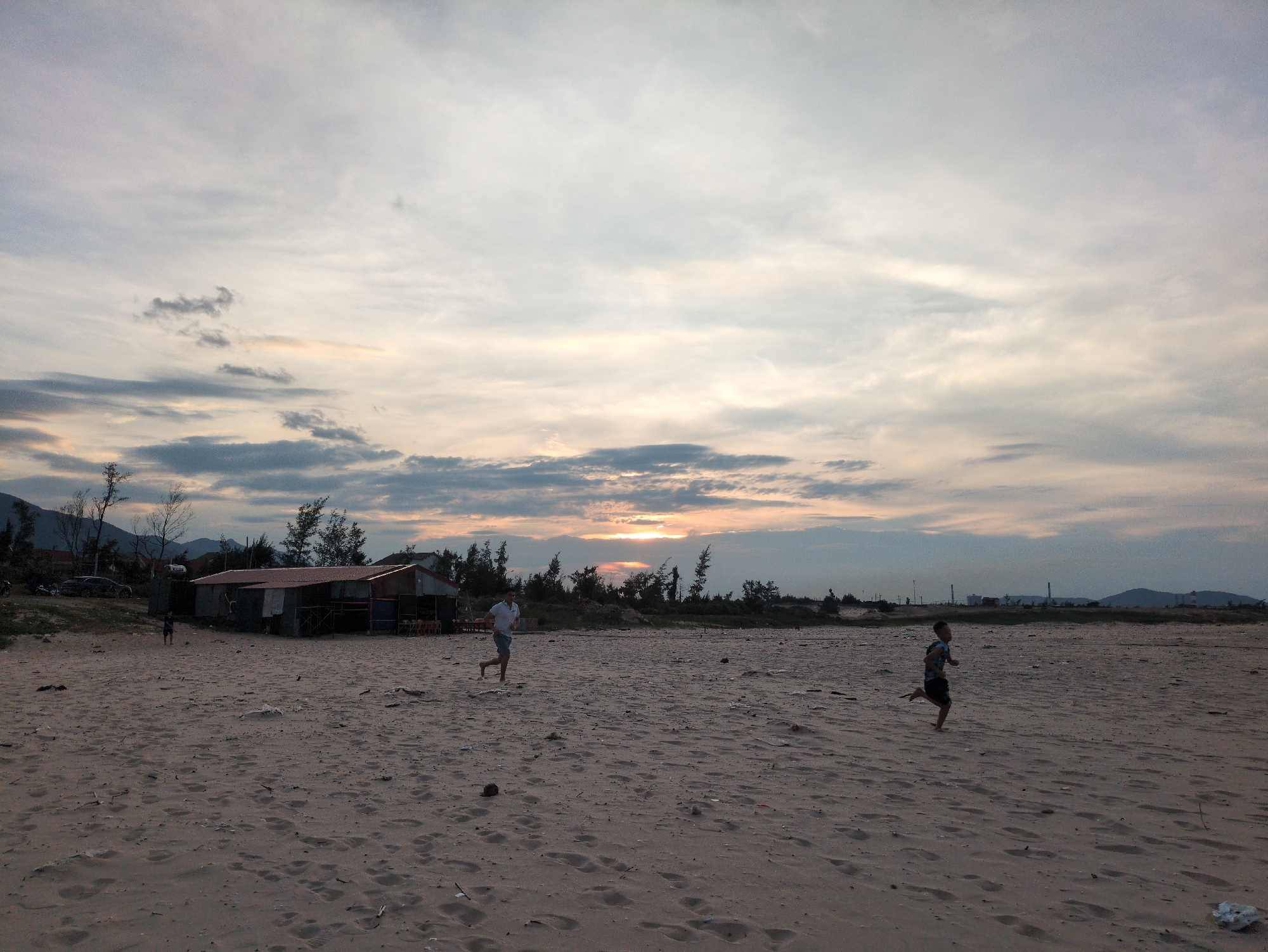 A wide-angle, low-light photograph depicts a sandy beach in Hoành Sơn Ward, Vietnam, at dusk. The sky is dominated by dramatic clouds, with a sliver of the setting sun visible on the horizon, casting a warm glow through the atmospheric haze. In the mid-ground, a small, rustic building with a corrugated metal roof and open sides stands on the sand, flanked by sparse trees. A few people are present on the beach. To the left of the building, a dark-clothed figure is visible. Further out in the sandy expanse, a man in a white shirt and blue shorts is captured mid-stride, running to the left. To the right, a young boy in dark shorts and a patterned shirt is also running to the left. The beach itself is uneven and textured with sand ripples and scattered debris. In the background, a line of trees separates the beach from distant hills and what appear to be industrial structures. The overall impression is one of a serene, yet active, coastal scene at the close of day.