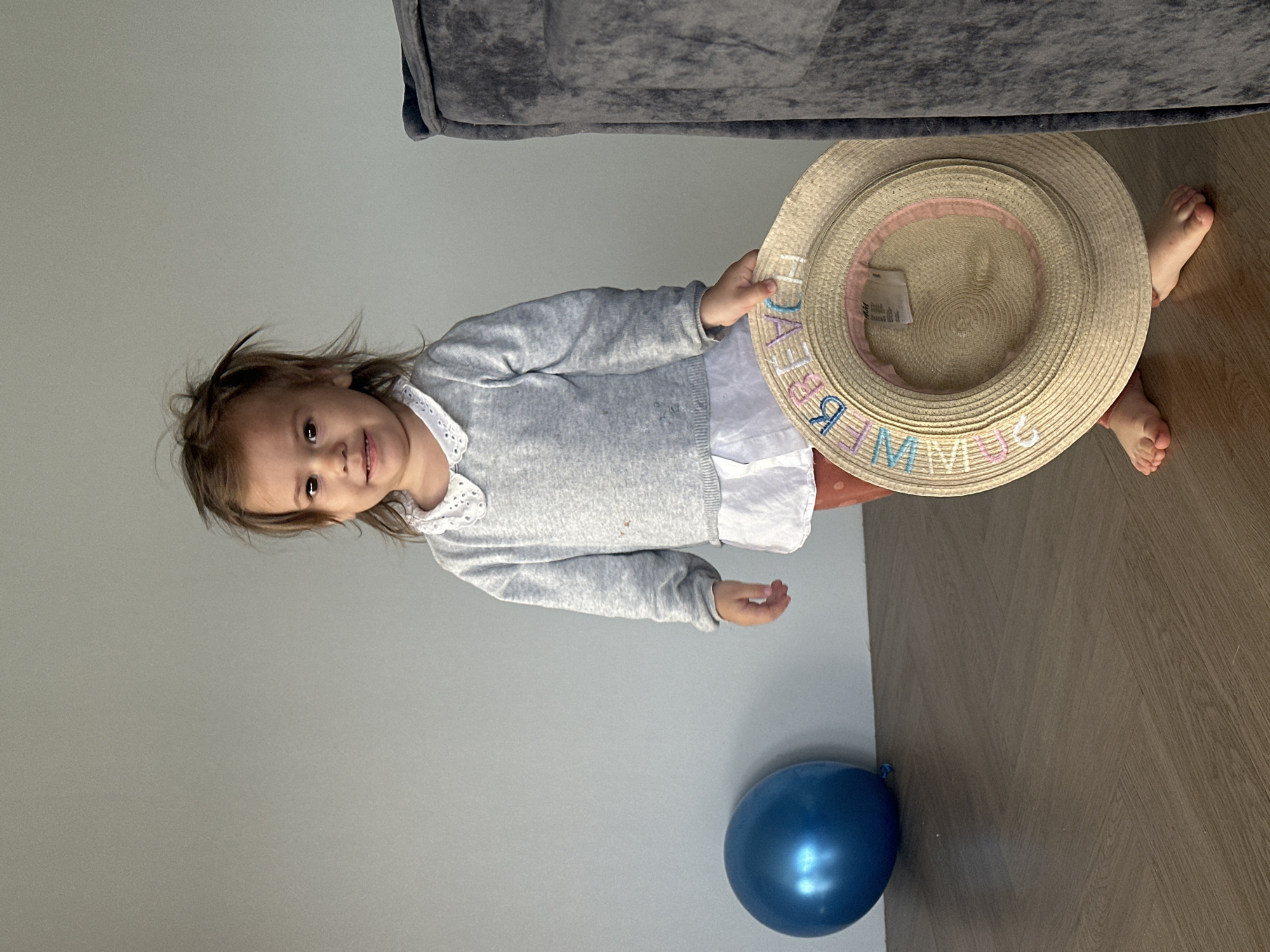 A full-body shot of a young child, approximately a toddler, standing indoors in Kharnavy, Russia. The child is looking towards the camera with a gentle smile, their light brown hair slightly disheveled. They are dressed in a white collared shirt under a grey knitted cardigan, with a partially visible reddish-brown clothing item on their lower body. Their bare feet are visible on the floor.
The child is holding a straw hat in their right hand, which has the word "SUMMER" embroidered in colorful letters (blue, pink, orange, green, light blue, pink) around its brim. The background consists of a plain light gray wall and a dark wooden floor with a herringbone pattern. A shiny blue balloon rests on the floor to the left of the child. A dark gray sofa or armchair is partially visible in the upper background. The natural lighting suggests the scene takes place during the daytime.